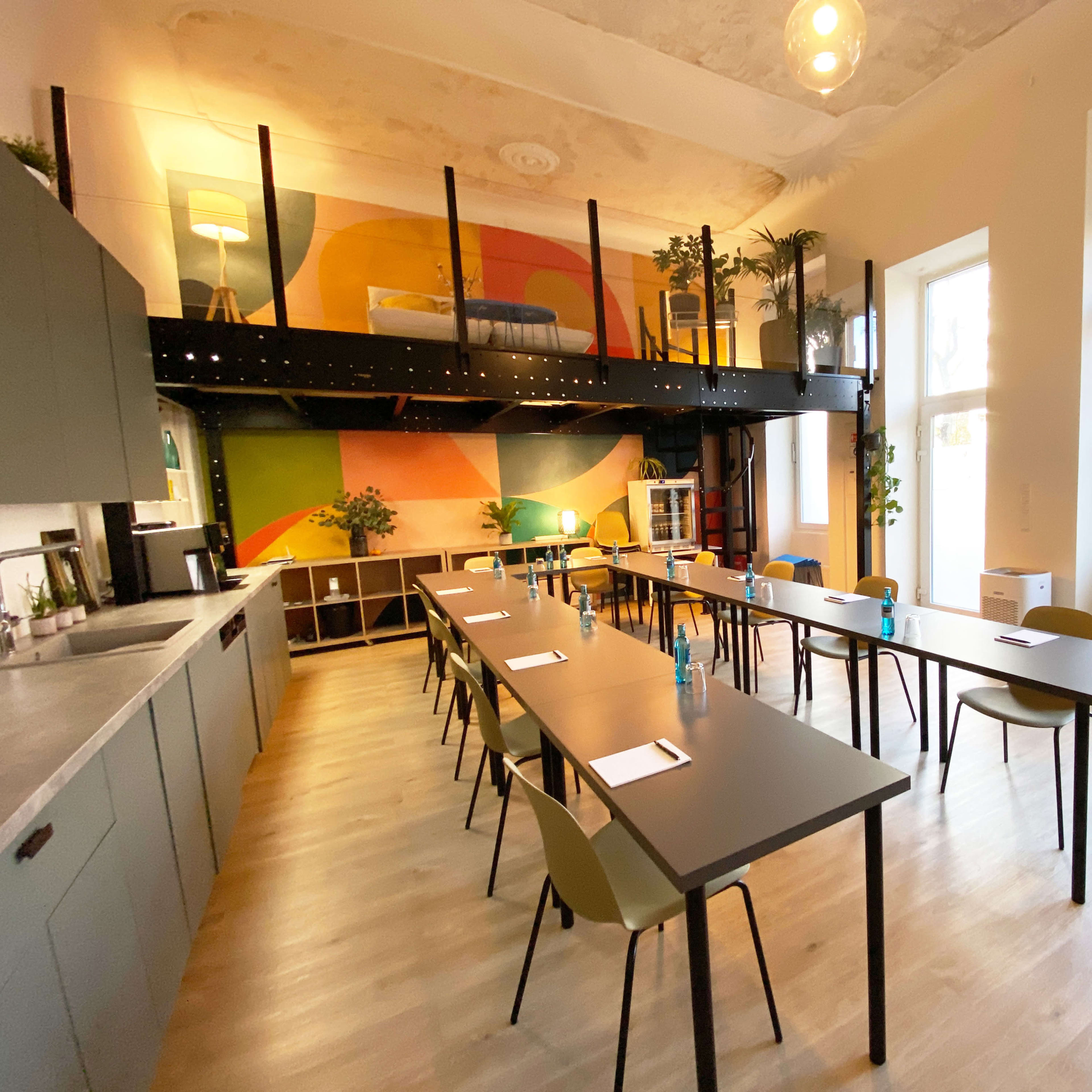 A spacious room features long tables arranged in rows, with a kitchenette and colorful wall art, illuminated by natural light from large windows.