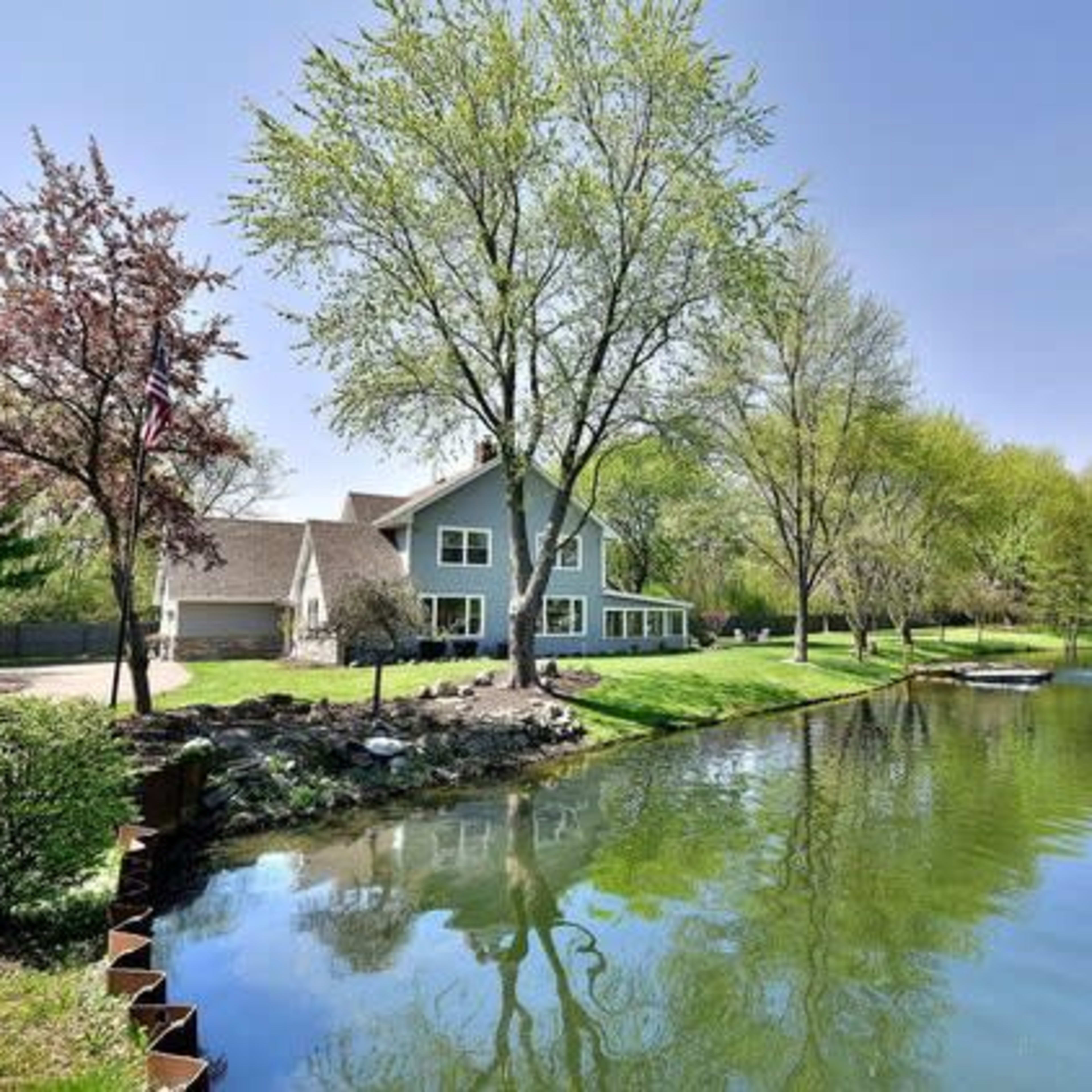 A house with large windows stands near a calm pond surrounded by green trees and landscaping.