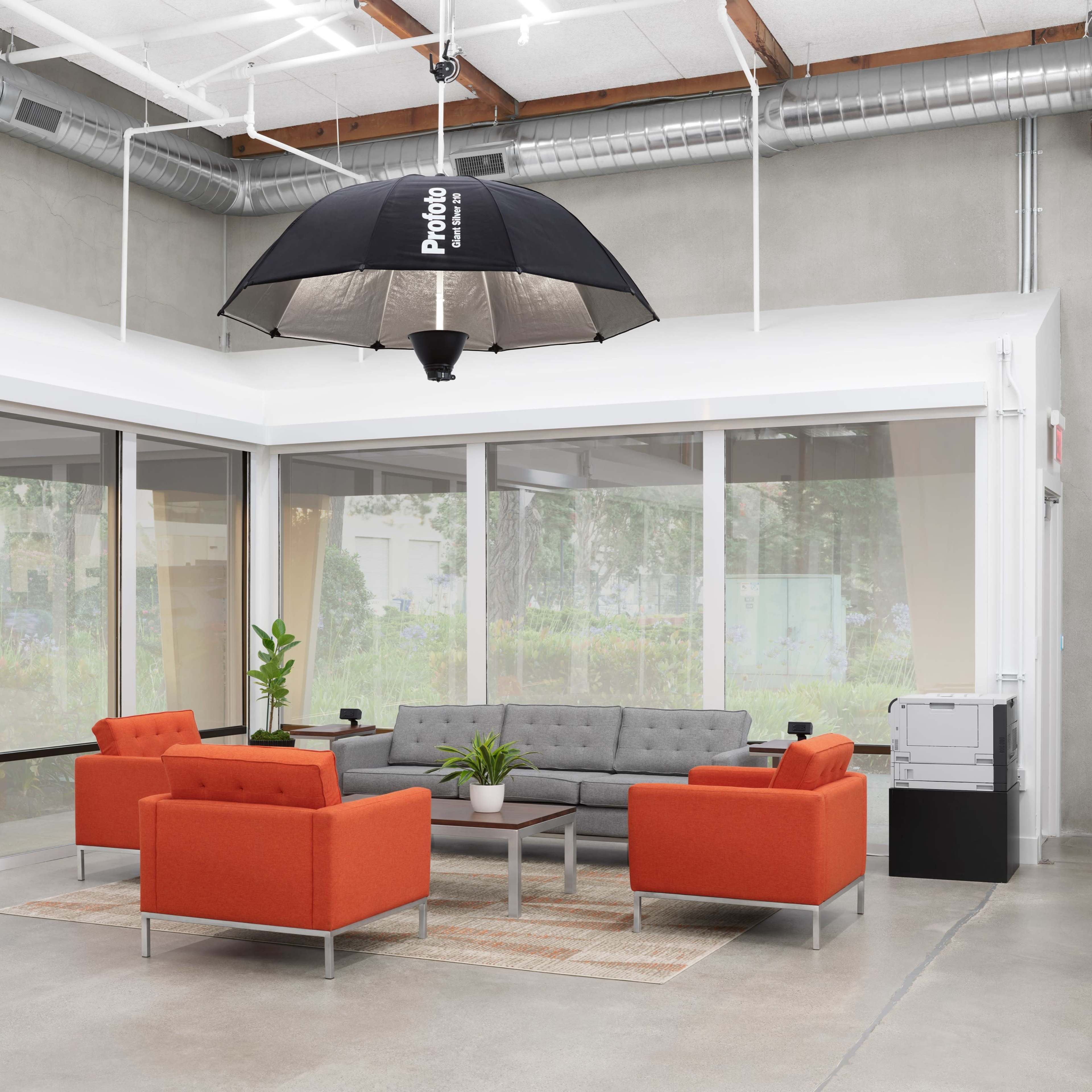 A modern lounge area with a gray sofa, two orange chairs, and a large umbrella light overhead, surrounded by windows.