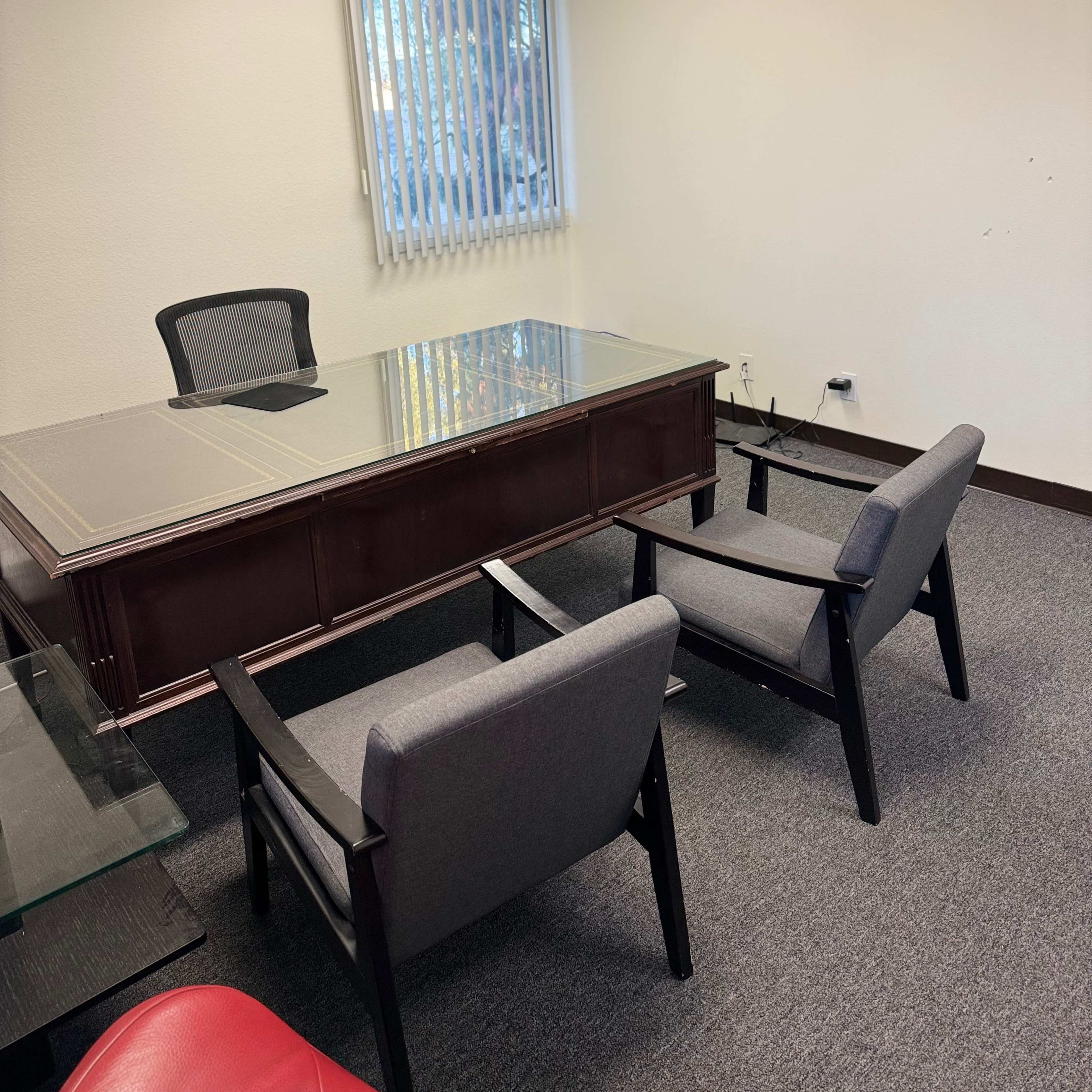A small office contains a wooden desk with a glass top and two gray chairs positioned in front of it, along with a window covered by vertical blinds.