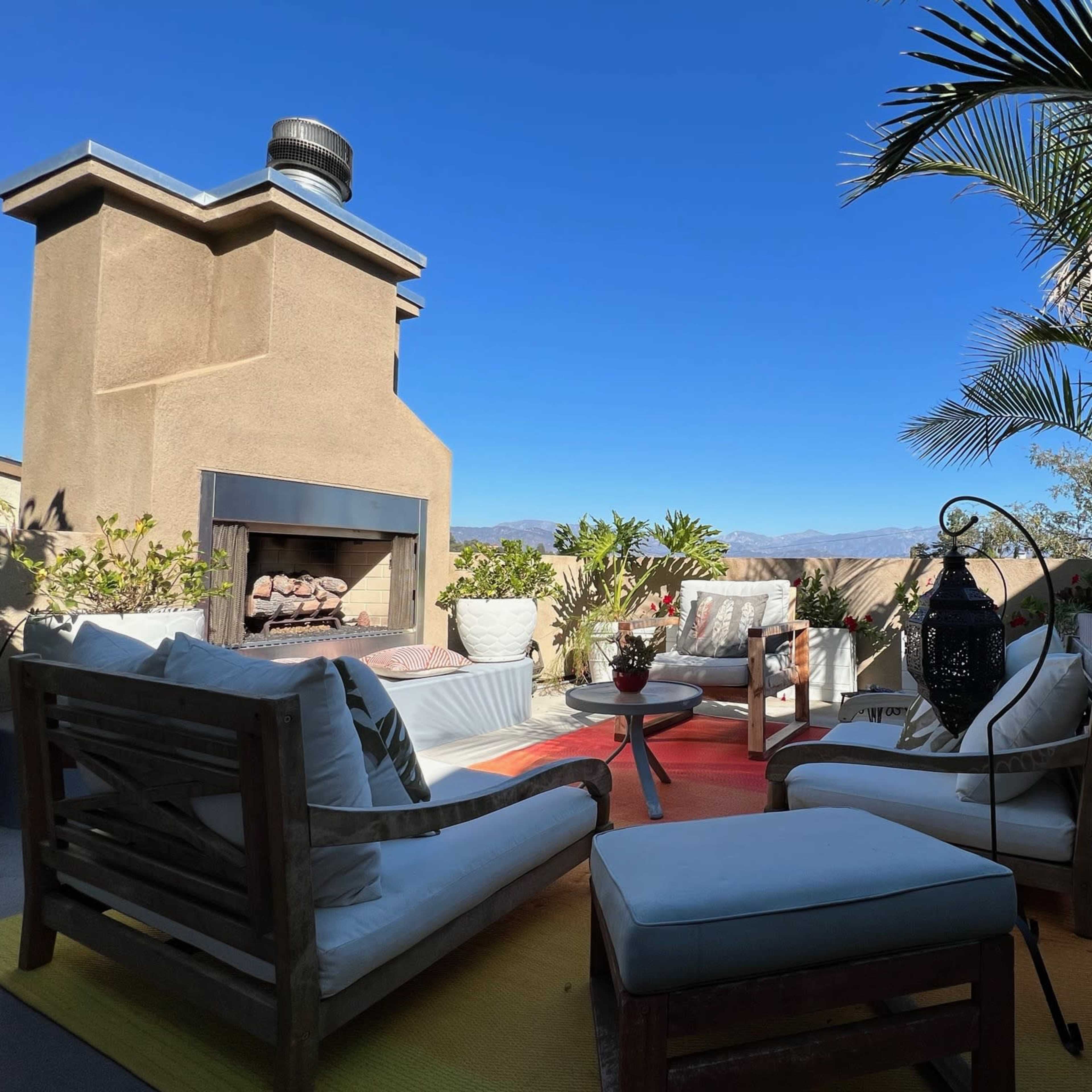 A rooftop patio with a seating area, featuring a fireplace and potted plants under a clear blue sky.