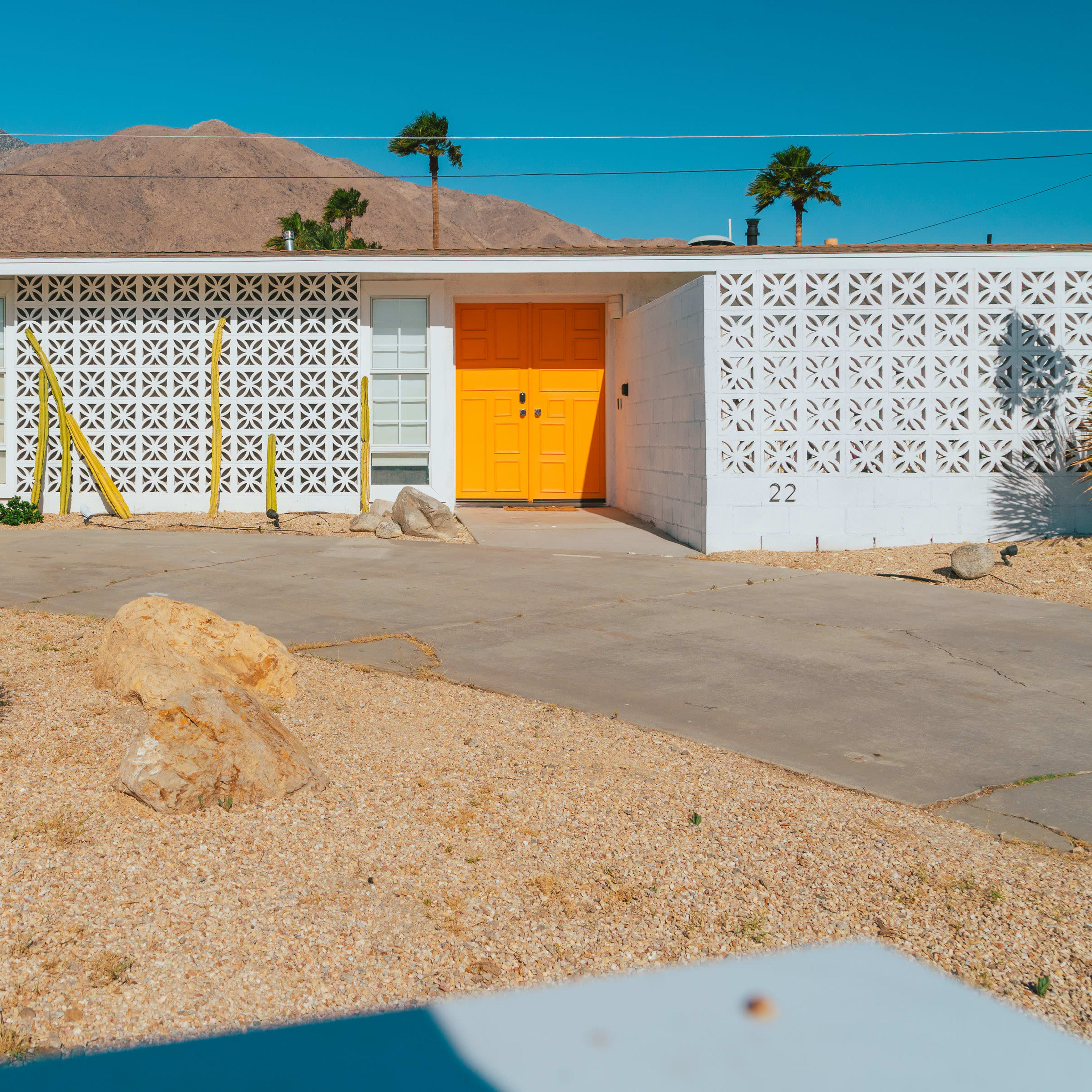 A single-story house with a bright orange door and a decorative wall stands in a desert landscape featuring palm trees and gravel.