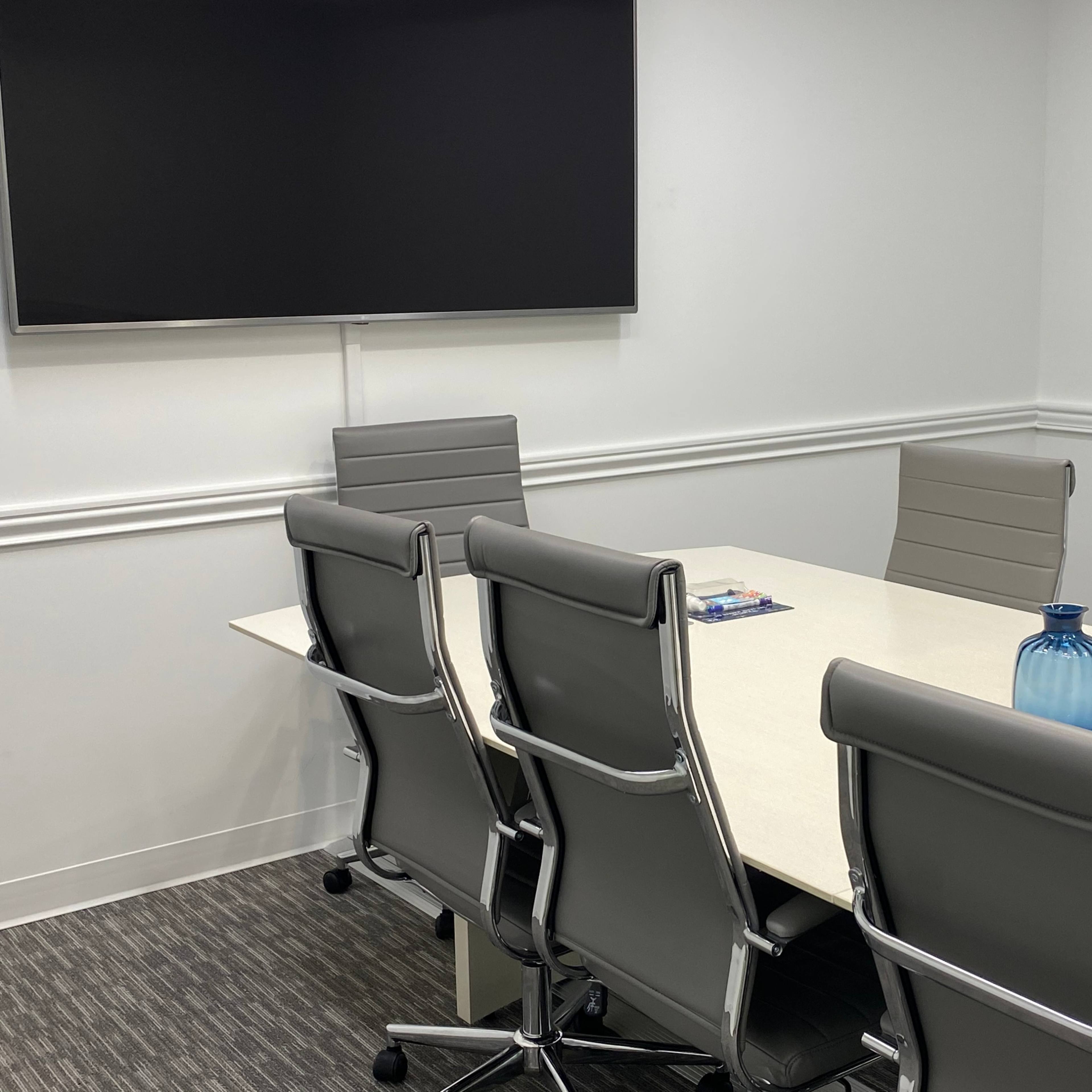 A conference room features a long table with six gray chairs, a blue vase, and a wall-mounted television.