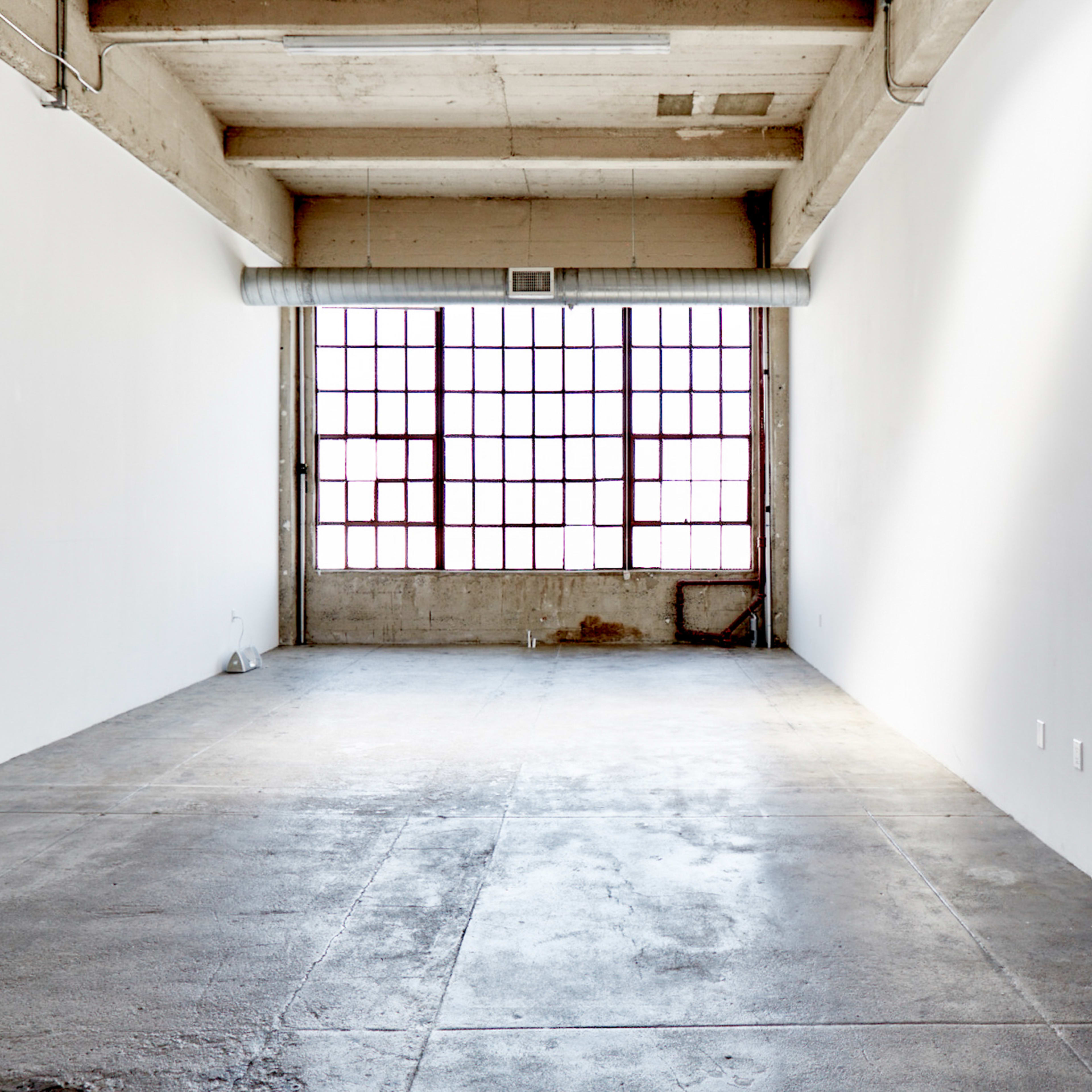 An empty room with concrete flooring and large windows along one wall, allowing natural light to enter.