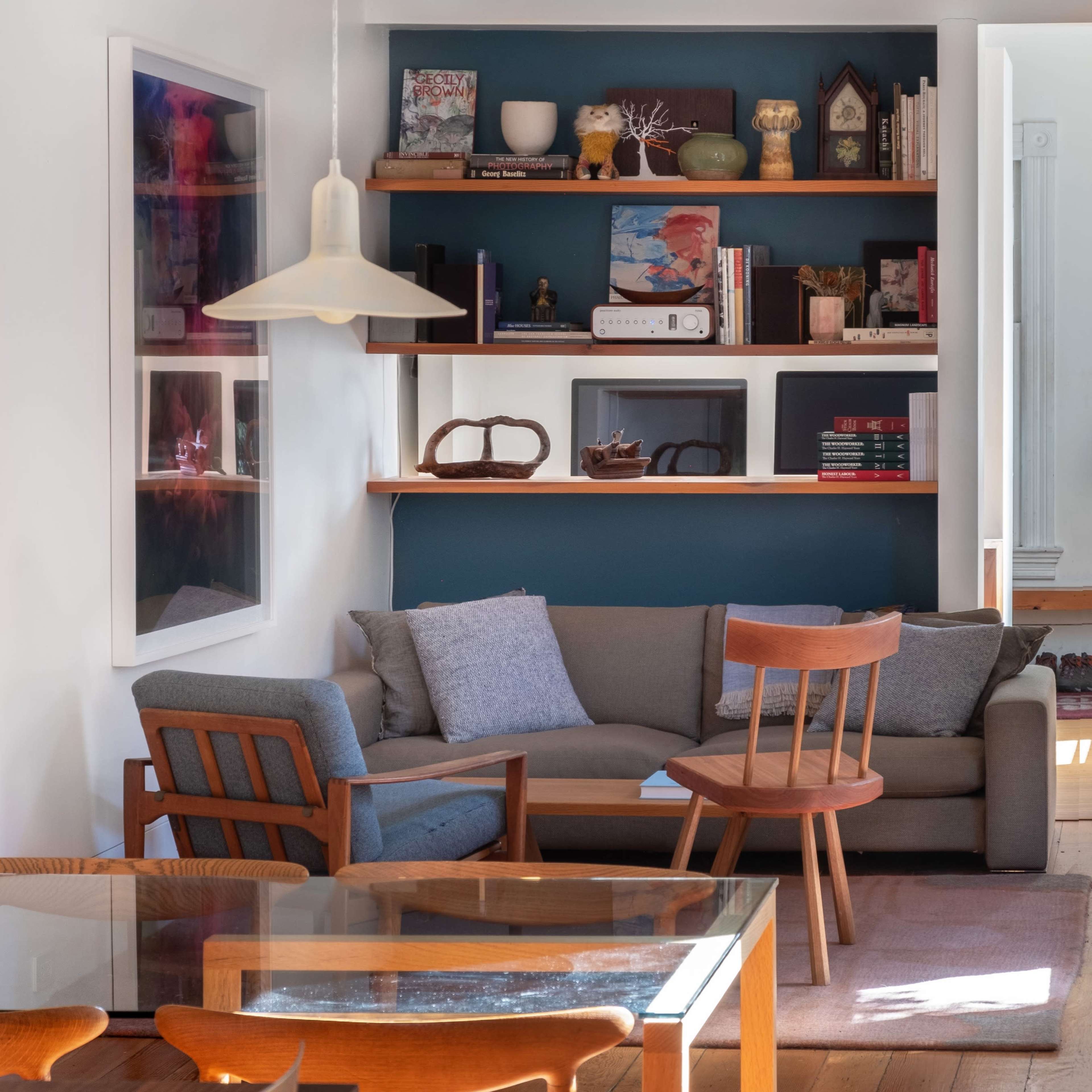 A living space with a gray sofa, a wooden chair, a dining table, and shelves displaying various books and decorative items against a blue wall.
