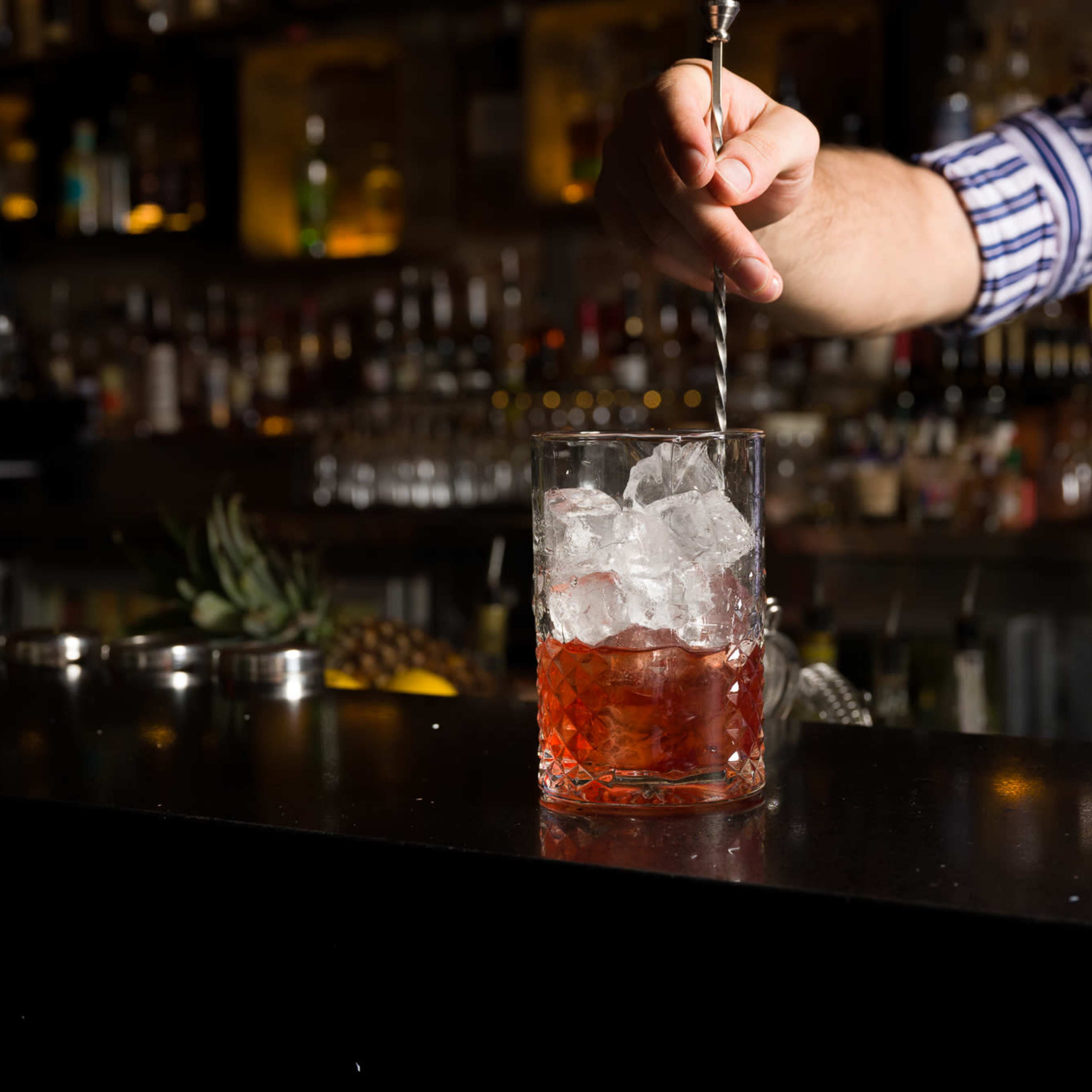 A bartender stirs a cocktail in a glass filled with ice at a bar.