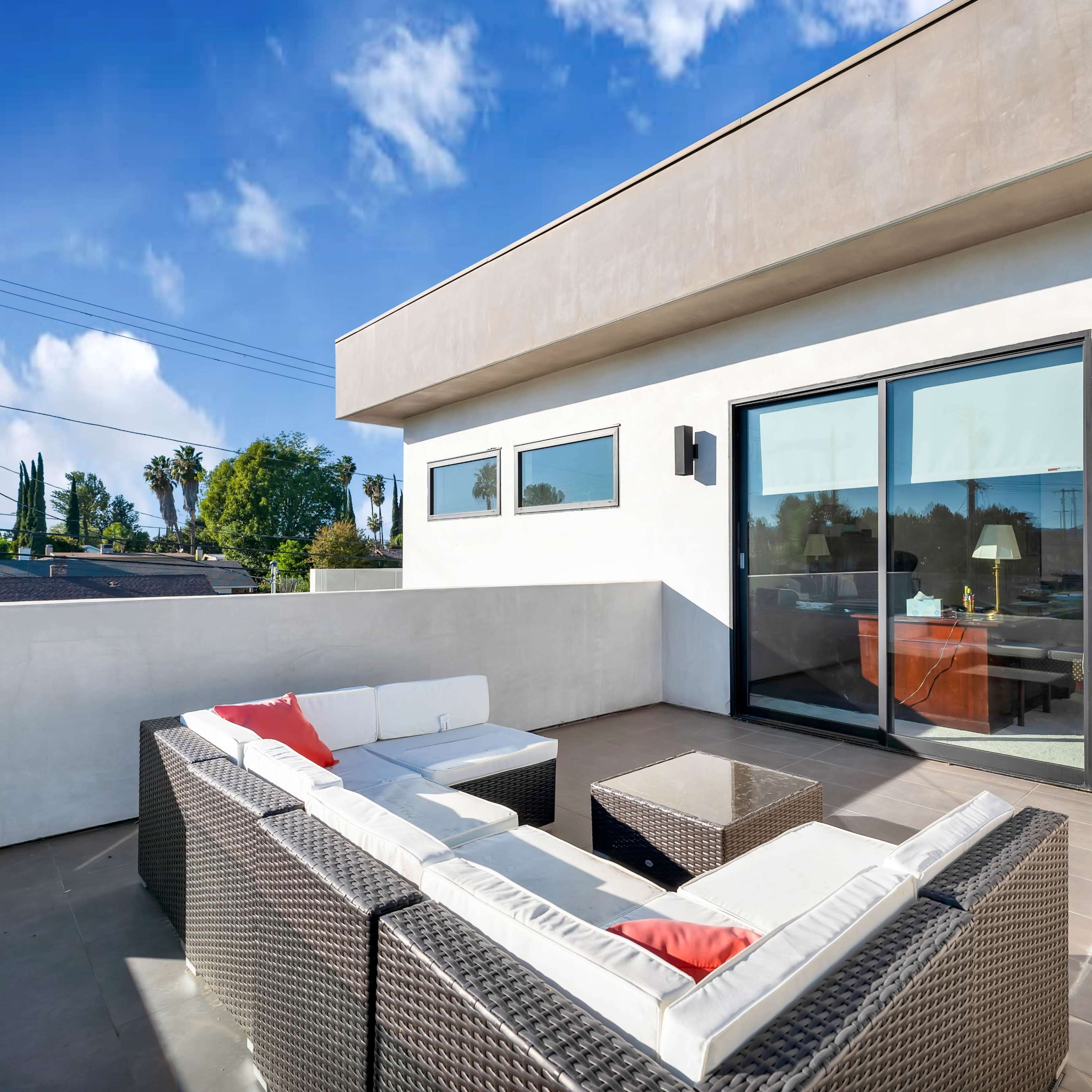 A modern outdoor living space features a sectional sofa with cushions, a coffee table, and large glass doors leading indoors.