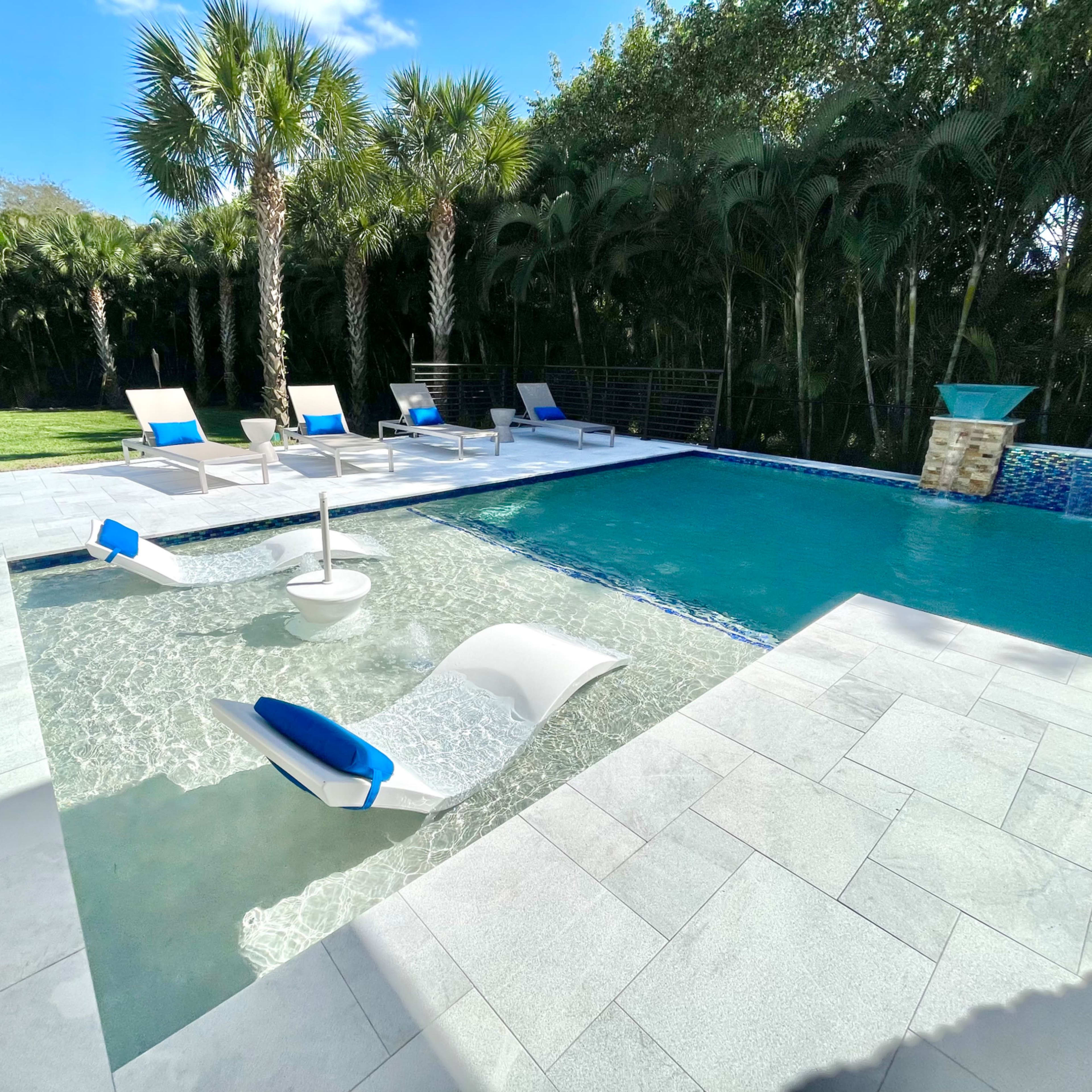 A modern outdoor pool area with several lounge chairs placed around the water, surrounded by palm trees and green landscaping.