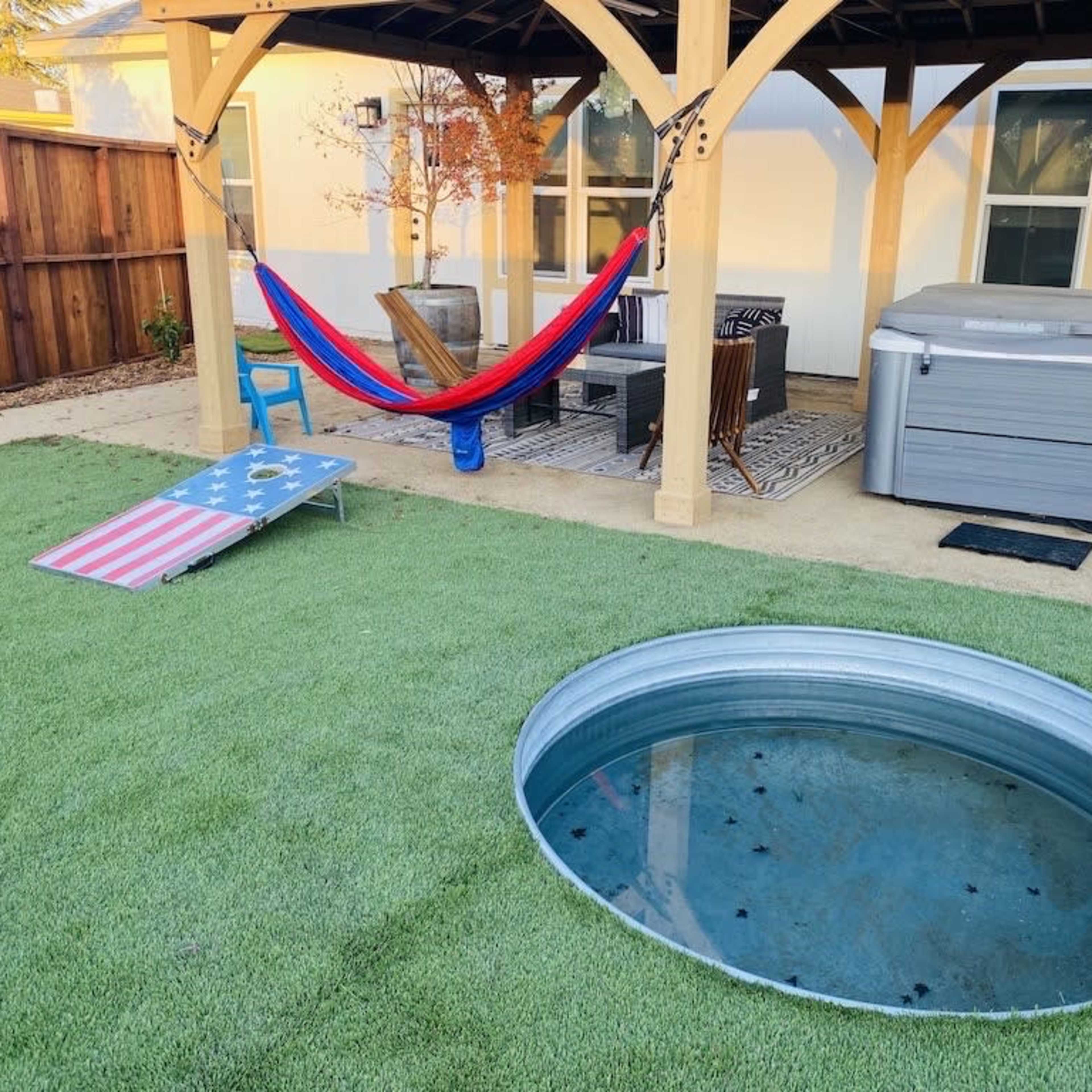A backyard with a hammock under a wooden pergola, a blue chair, a cornhole board featuring an American flag, a hot tub, and a circular metal tank filled with water on synthetic grass.