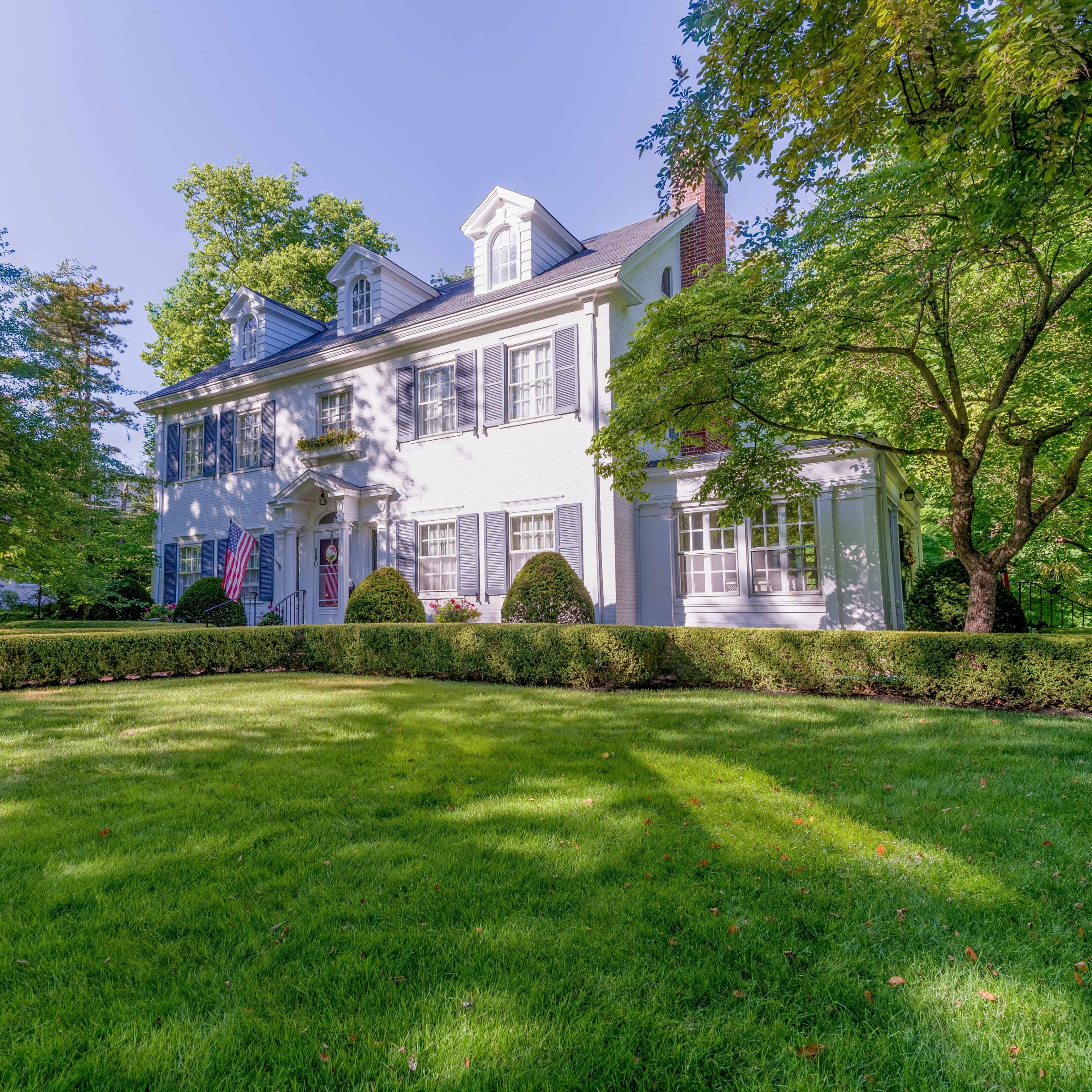 A white two-story house surrounded by well-maintained greenery and shrubs in a residential area.