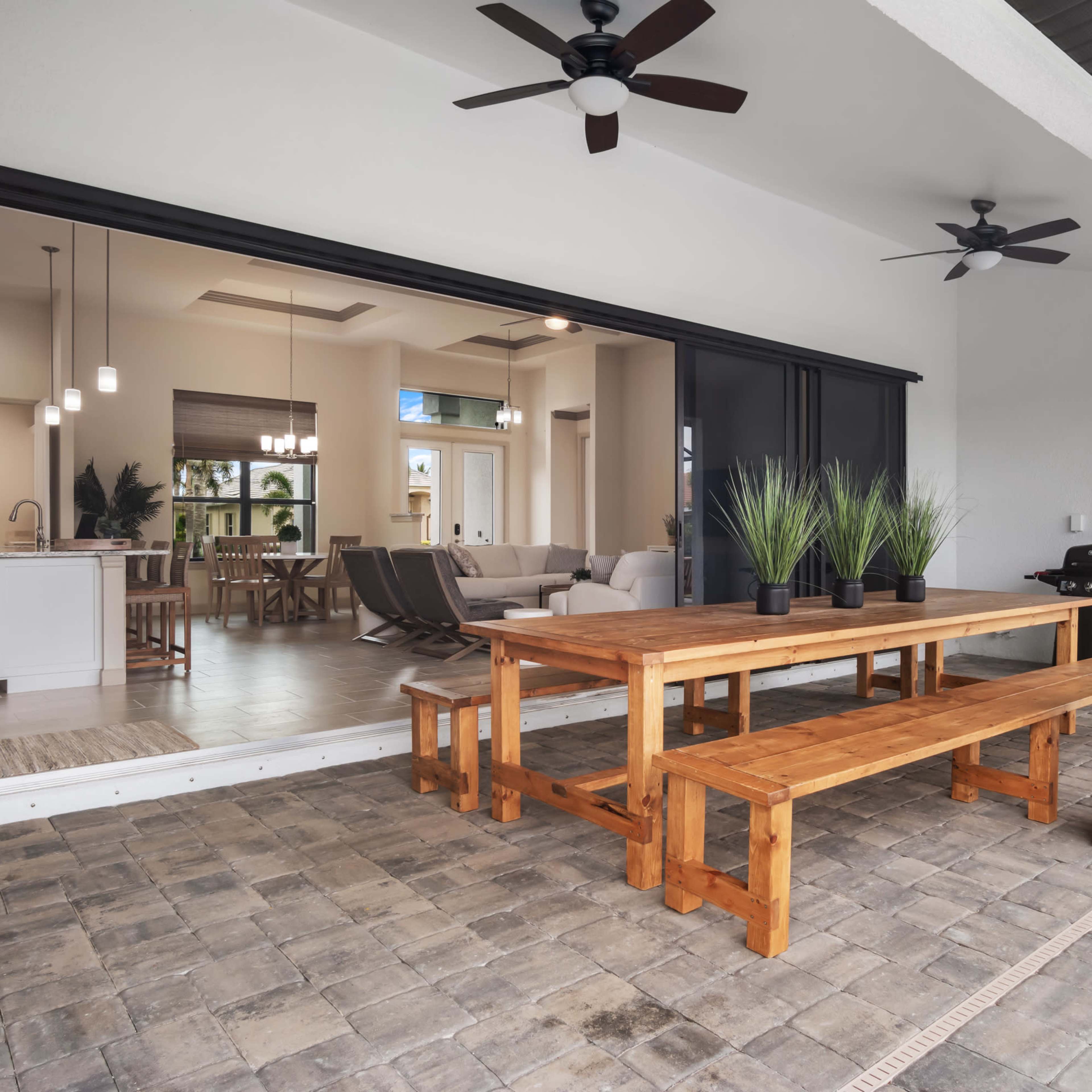 A wooden dining table and benches are positioned on a patio adjacent to an open interior space featuring modern furnishings.