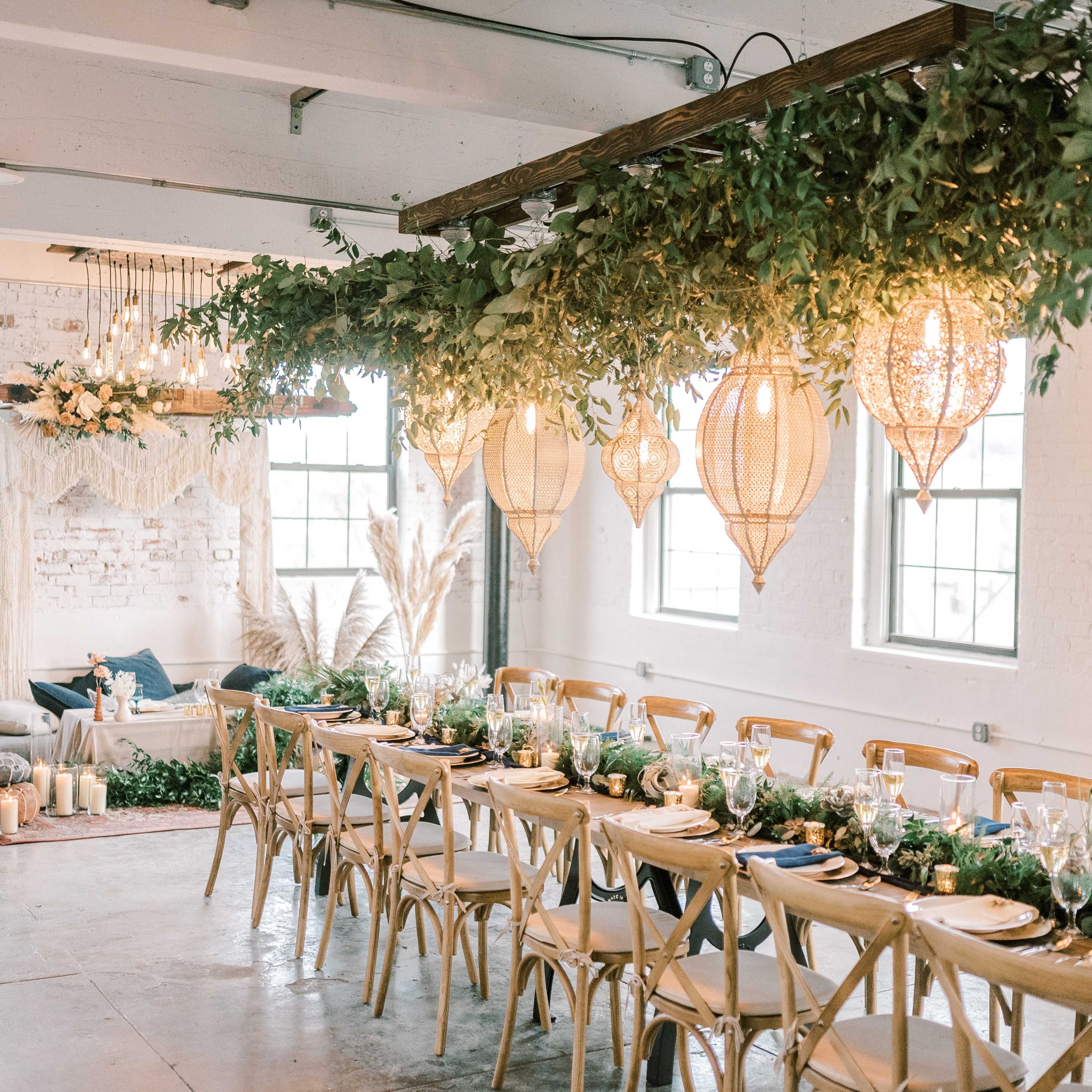 A decorated dining area featuring a long table with elegant table settings, surrounded by greenery and hanging lanterns, in a bright, industrial-style room with large windows.