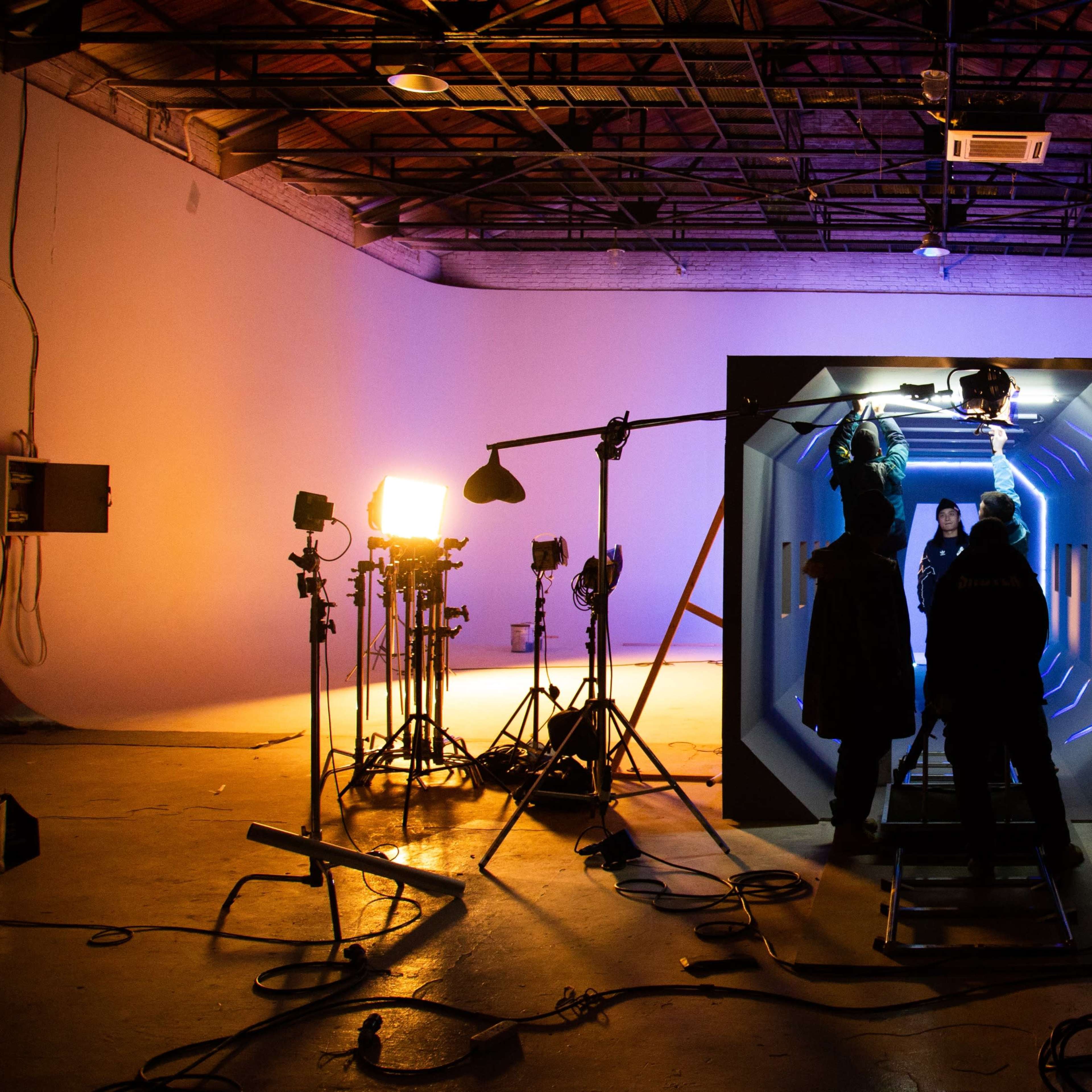 A group of people stands in a studio featuring a large, illuminated set piece with various lighting equipment and a ladder in the background.