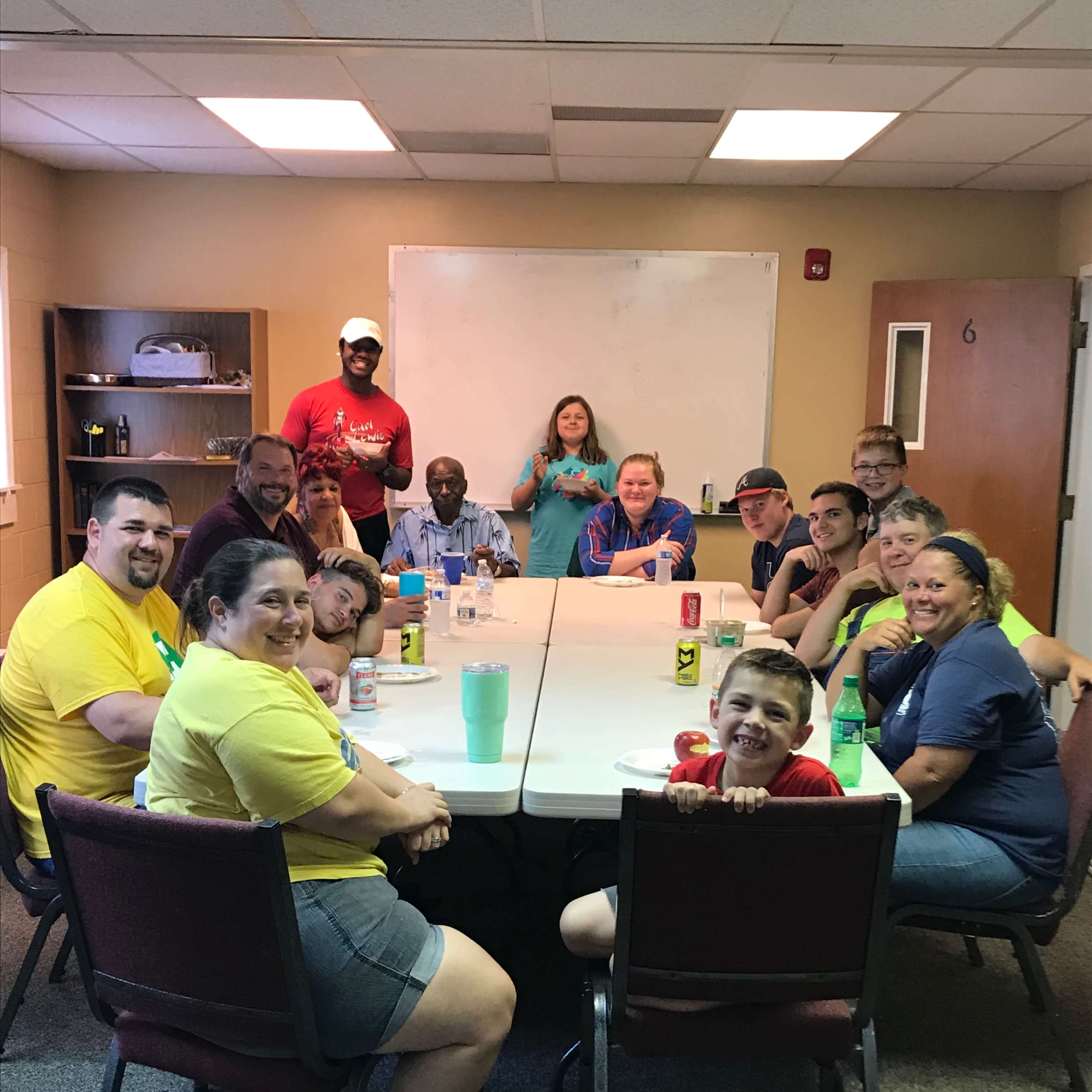 A diverse group of people sits around a long table in a room, all facing the camera while enjoying refreshments.