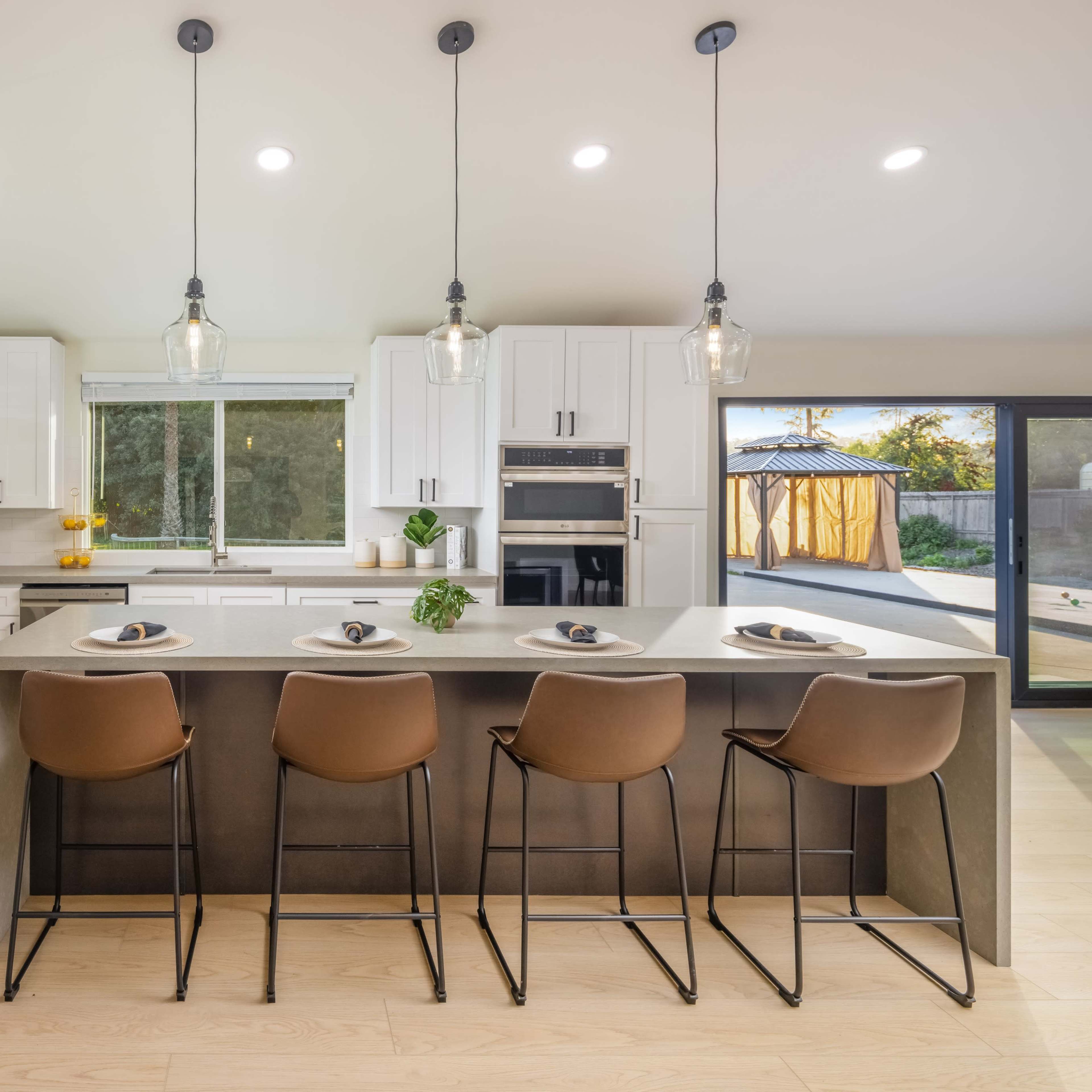 A modern kitchen features a concrete island with four brown bar stools, stainless steel appliances, and bright overhead lighting.