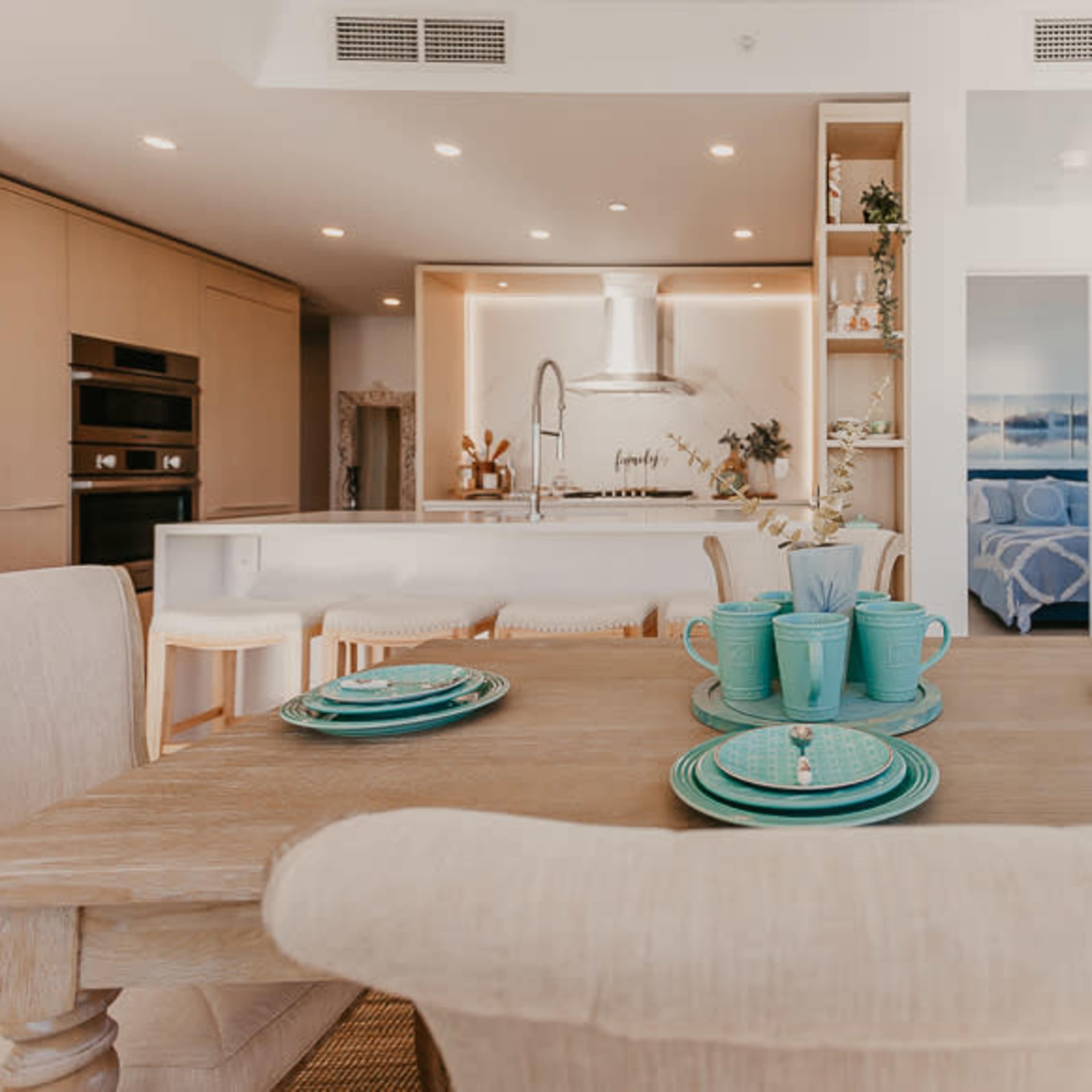 A dining area features a wooden table set with turquoise dishware, overlooking a modern kitchen with light wood cabinetry and a bright, airy design.