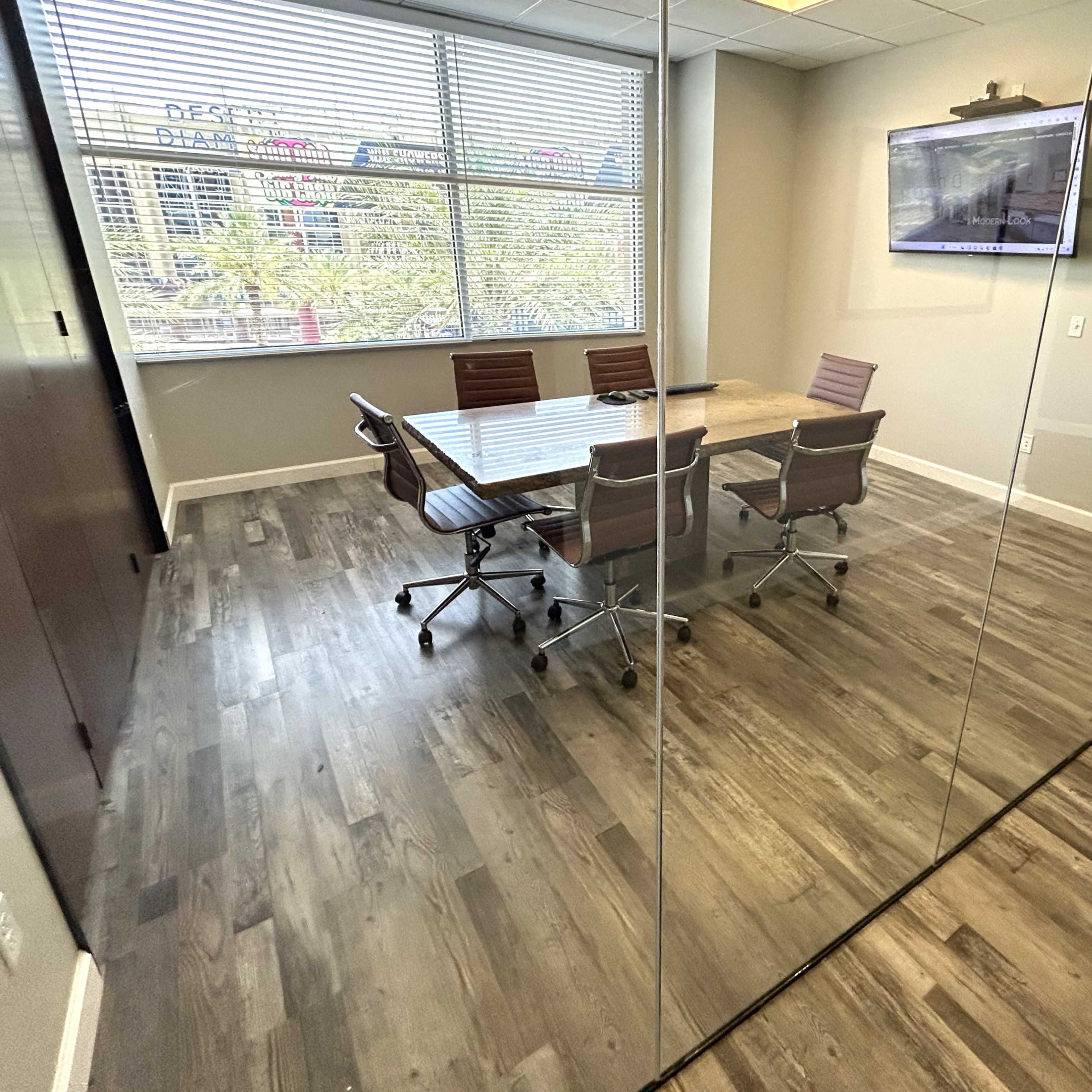 A modern conference room features a wooden table surrounded by brown leather chairs, with large windows offering a view of the outdoors.
