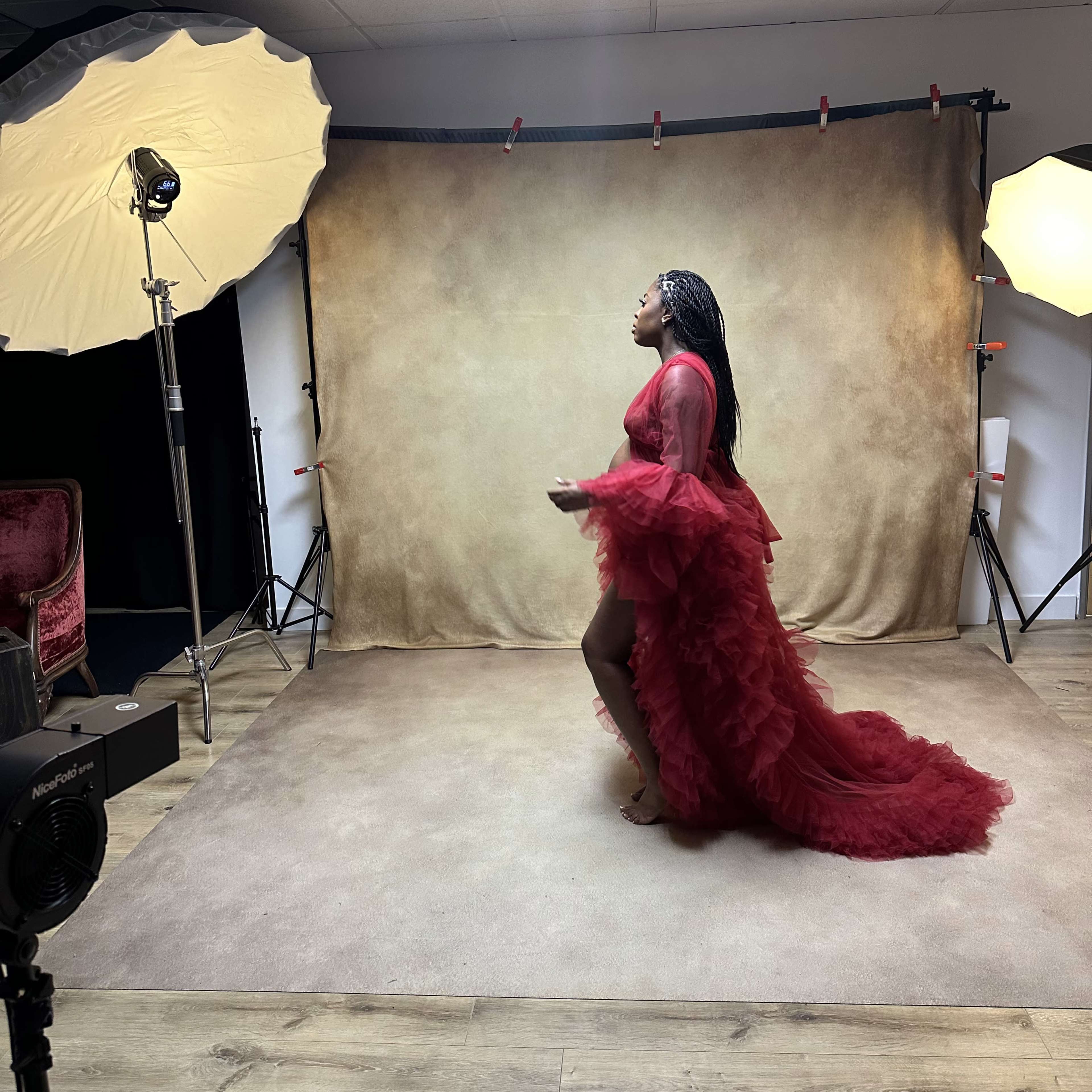A model in a red dress poses on a beige backdrop in a photography studio.