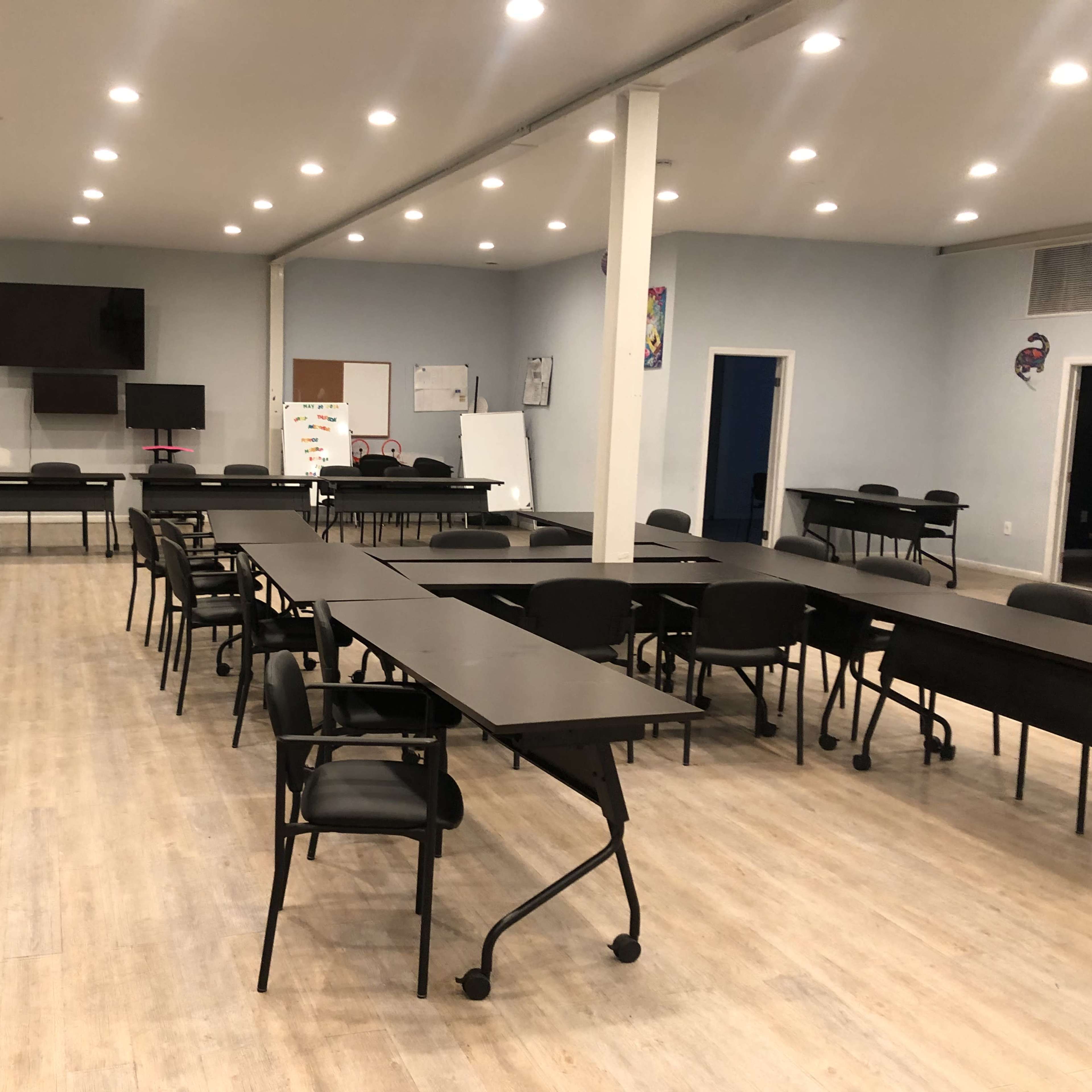 A spacious, empty classroom setup with several long black tables and chairs arranged in a professional layout, illuminated by bright ceiling lights.