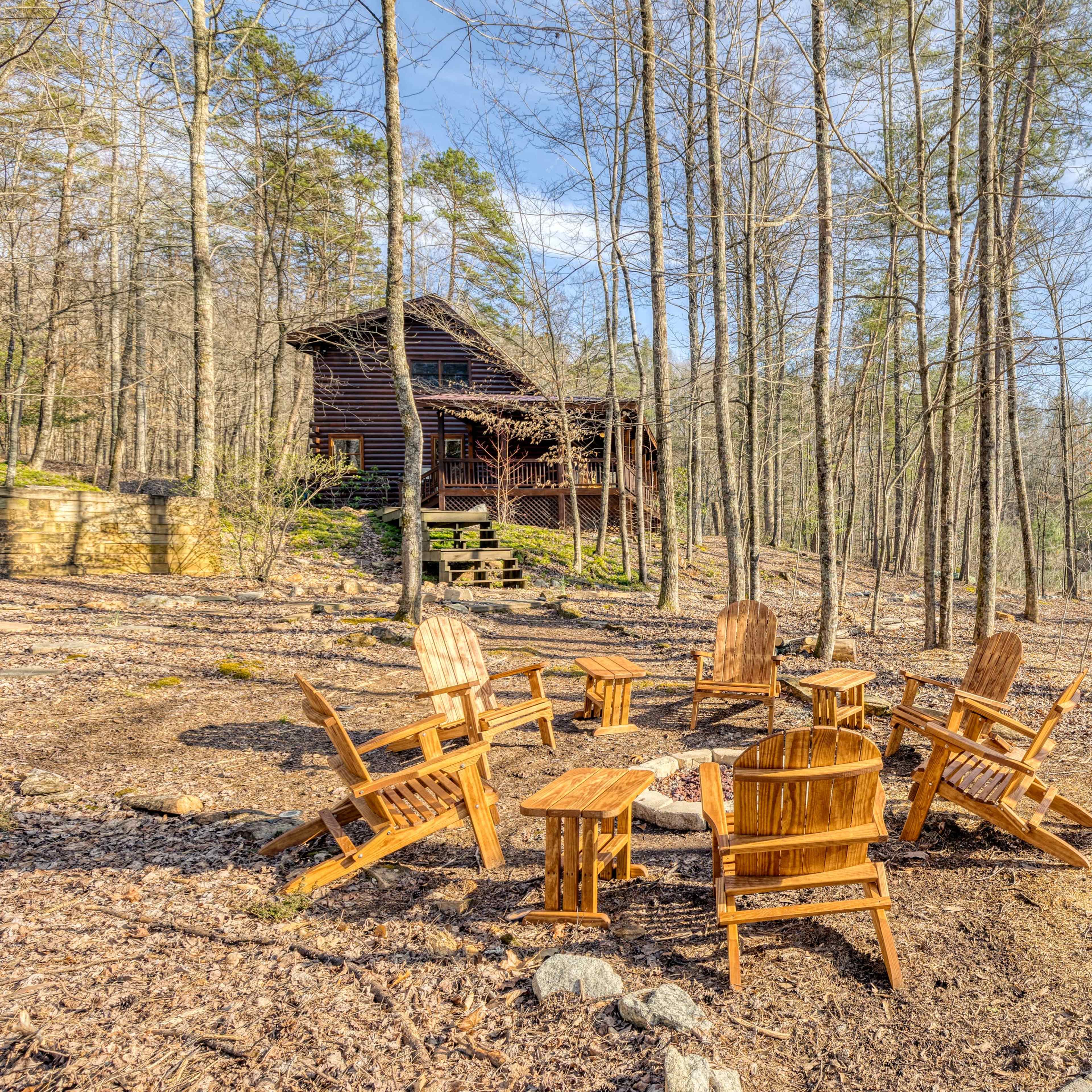 A circular seating area with wooden chairs and small tables is located in a forested area next to a lake, with a cabin visible in the background.
