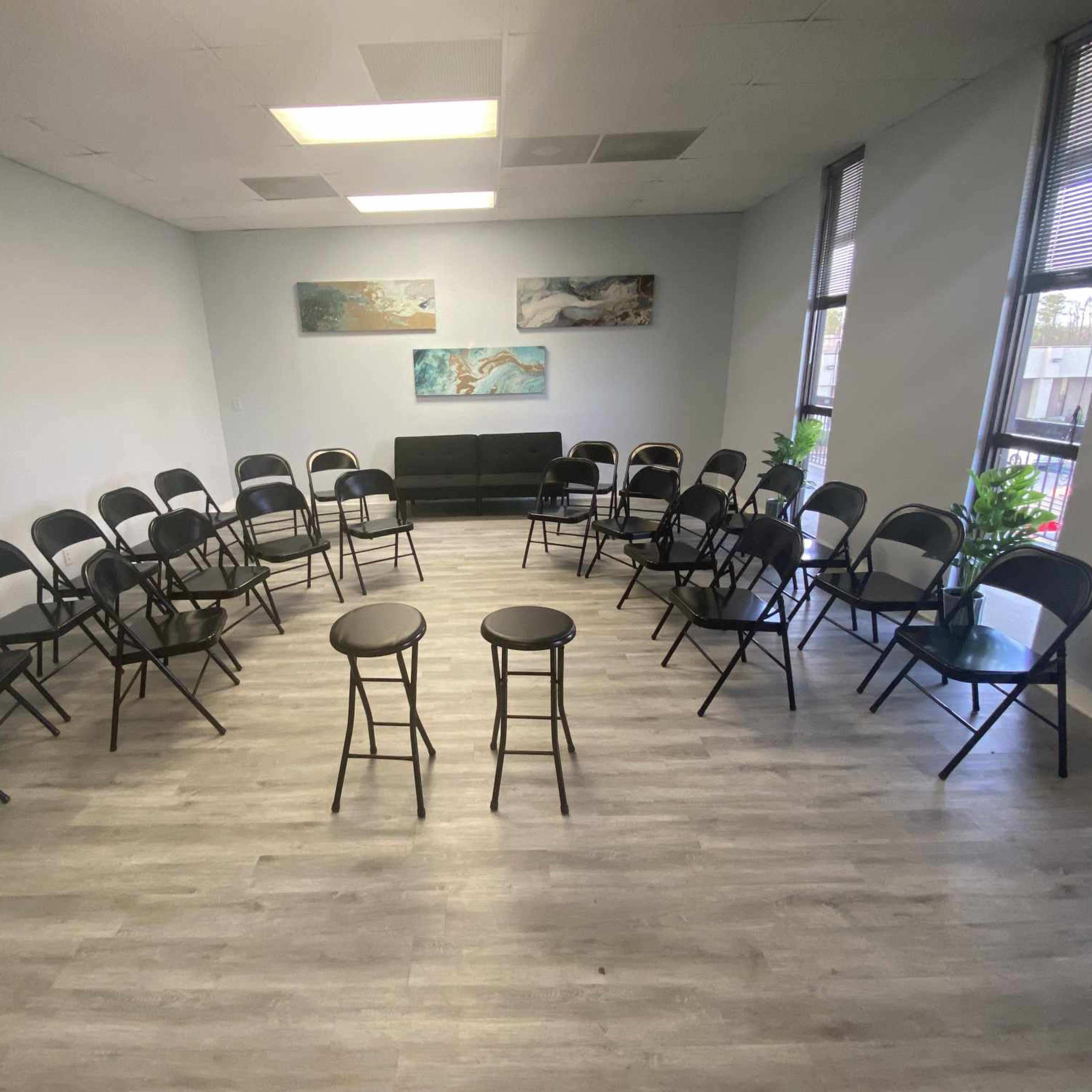 A circle of black folding chairs surrounds two tall stools in a well-lit room with gray walls and large windows.