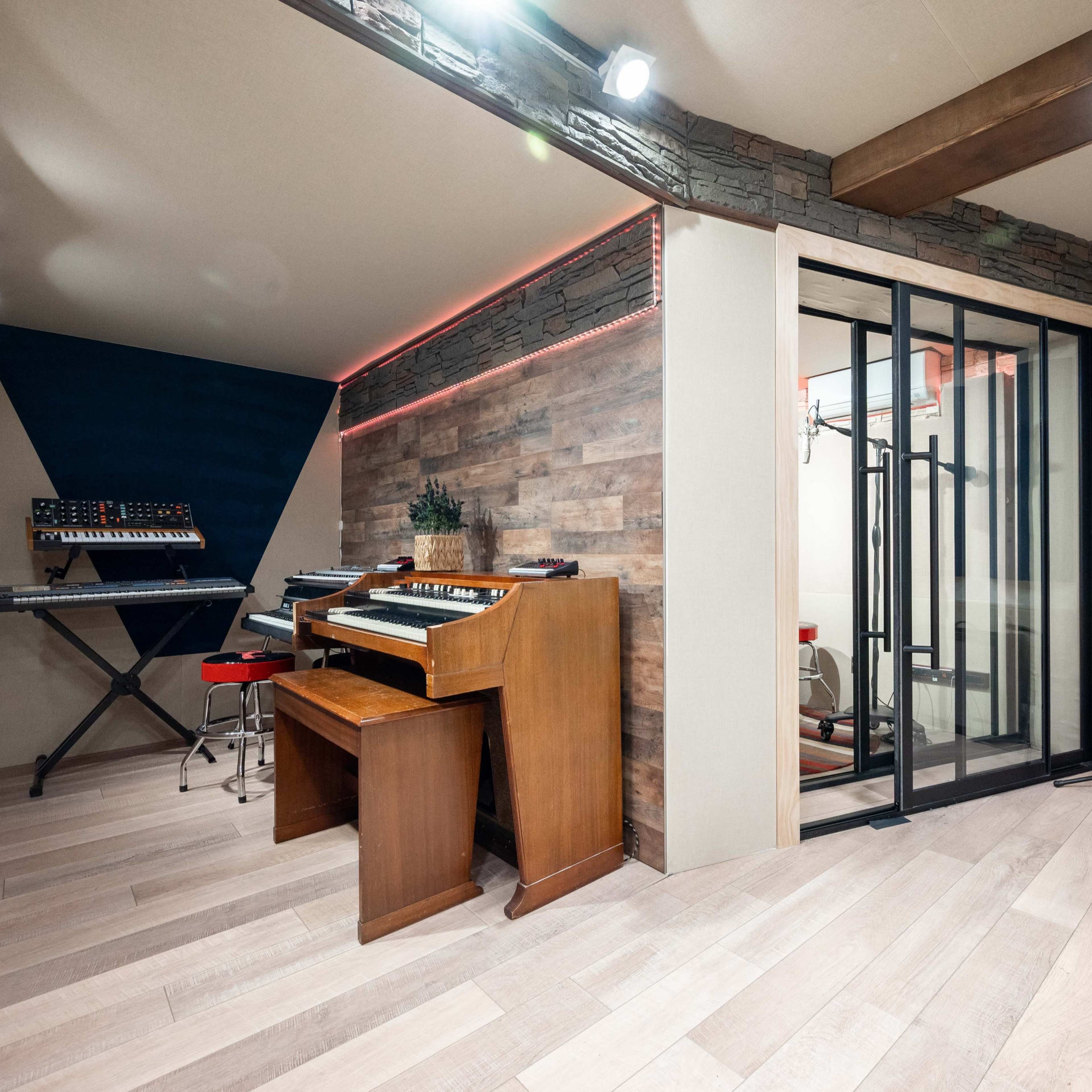 A modern music studio featuring a wooden keyboard, synthesizers, and soundproofing panels, with a glass enclosure for vocal recording.