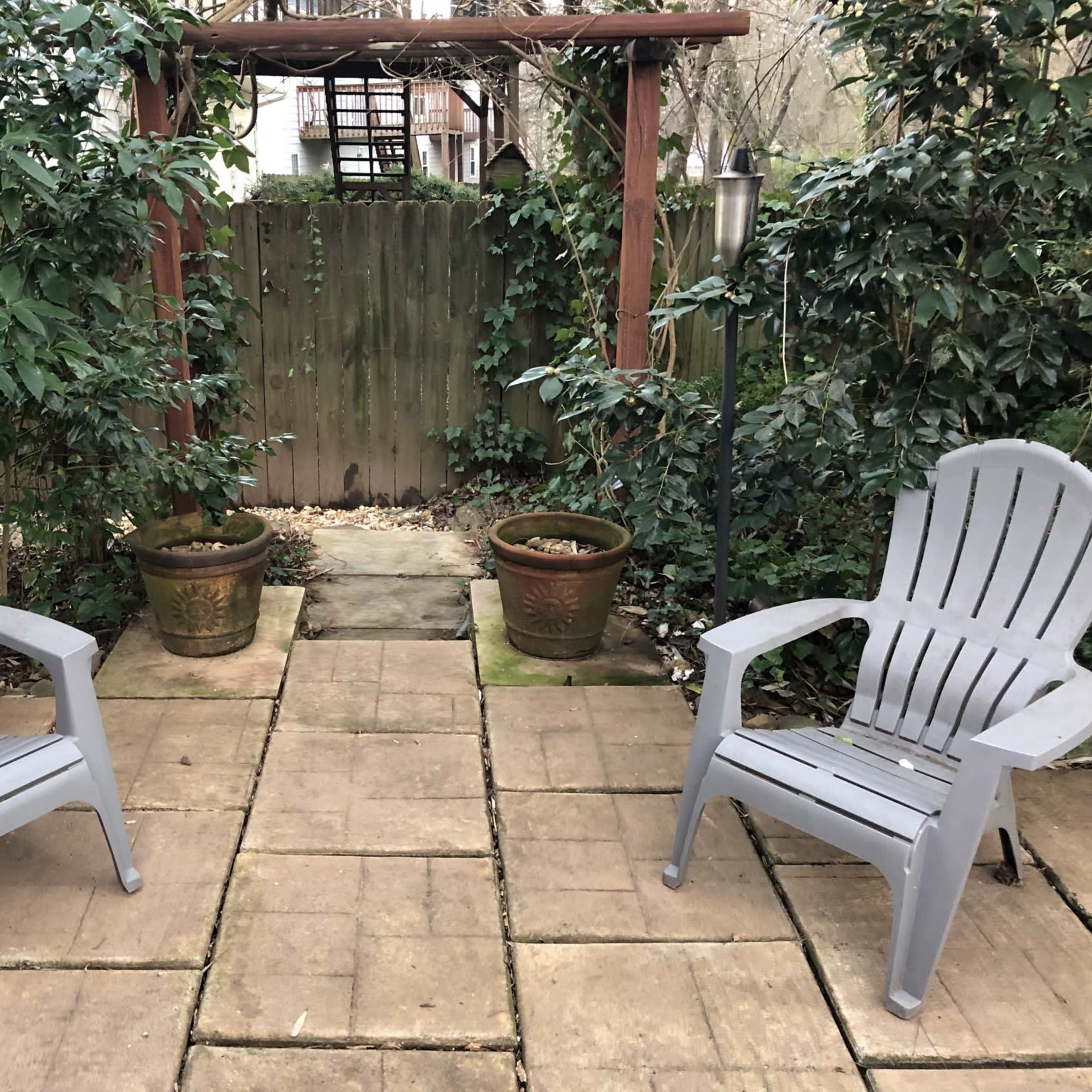 Two gray chairs are positioned on a stone patio surrounded by greenery and two potted plants.