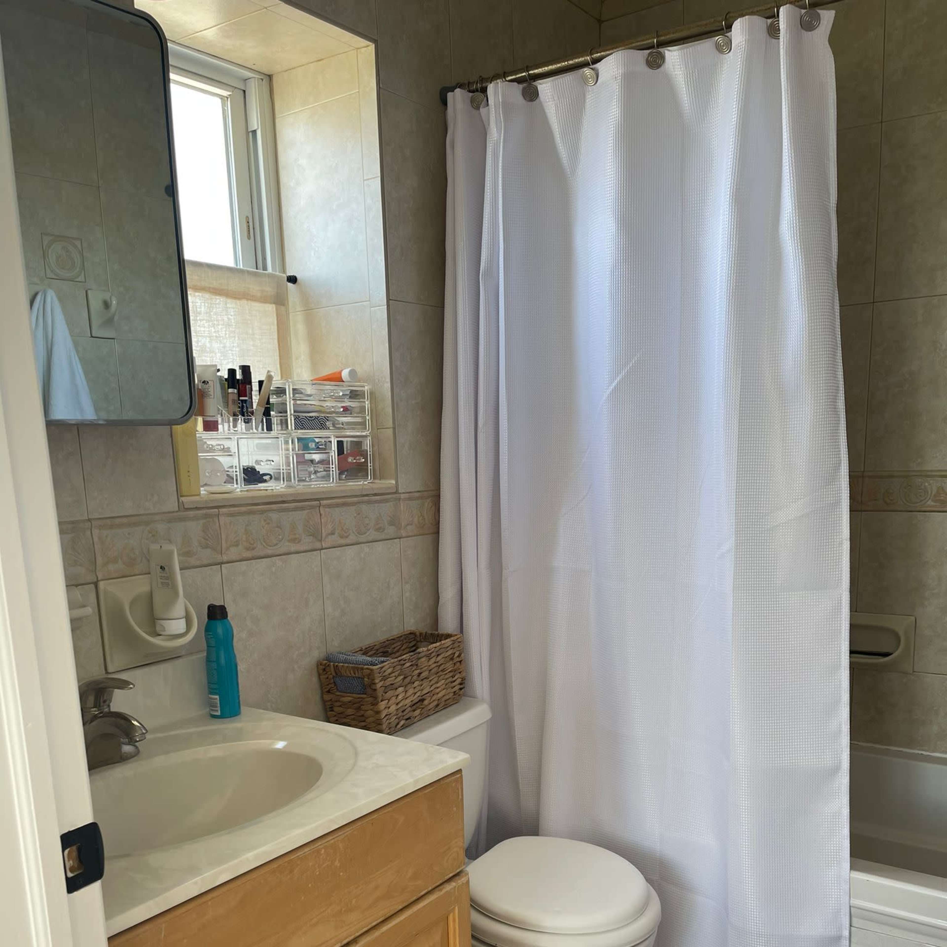 A bathroom with a sink and cabinet, a toilet, a white shower curtain, and a bathtub, all arranged around tiled walls and a window.