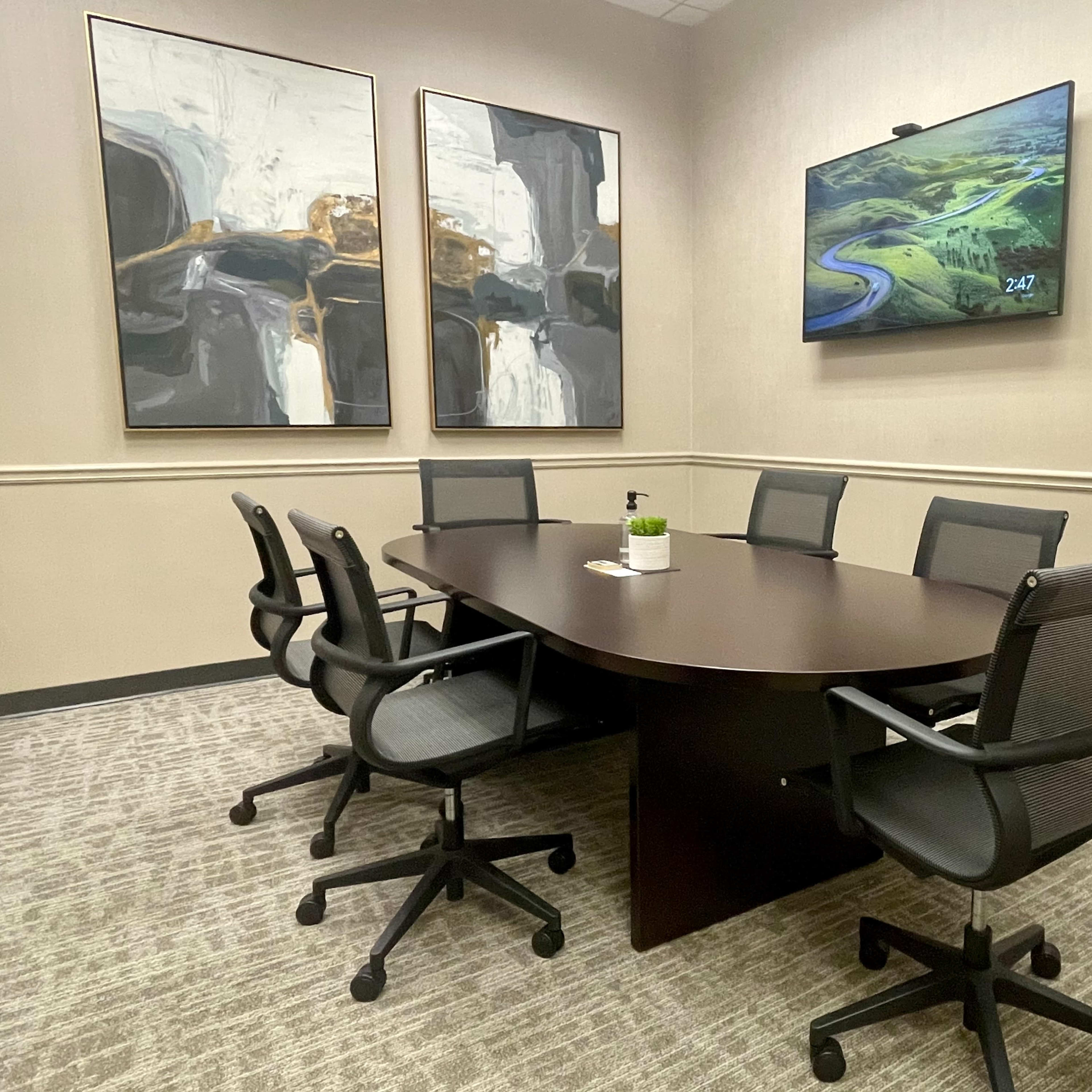 A conference room with a large oval table surrounded by six black office chairs, a television mounted on the wall, and abstract artwork on the adjacent walls.