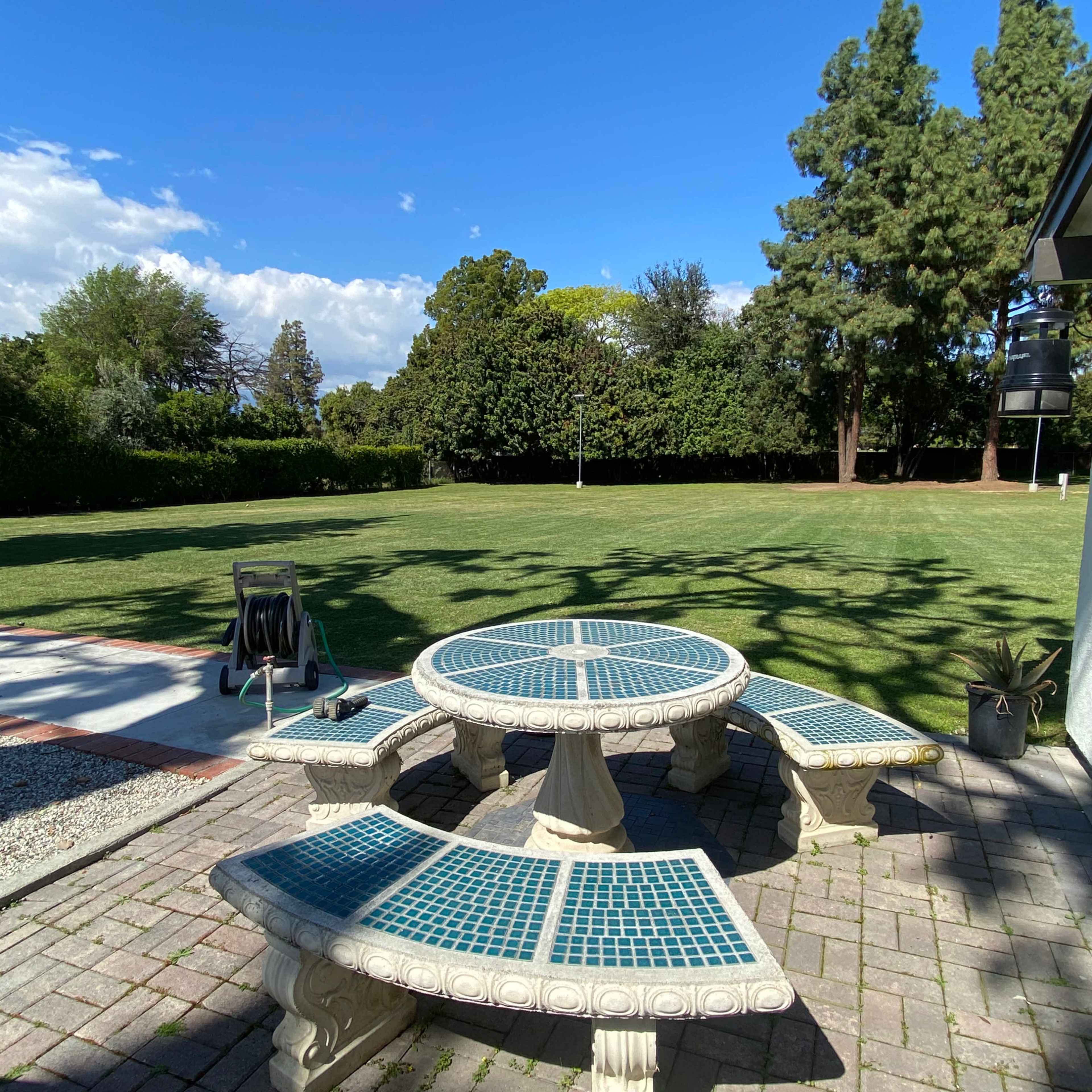 A stone table with tile accents and benches sits on a paved patio next to a large, grassy lawn surrounded by trees.