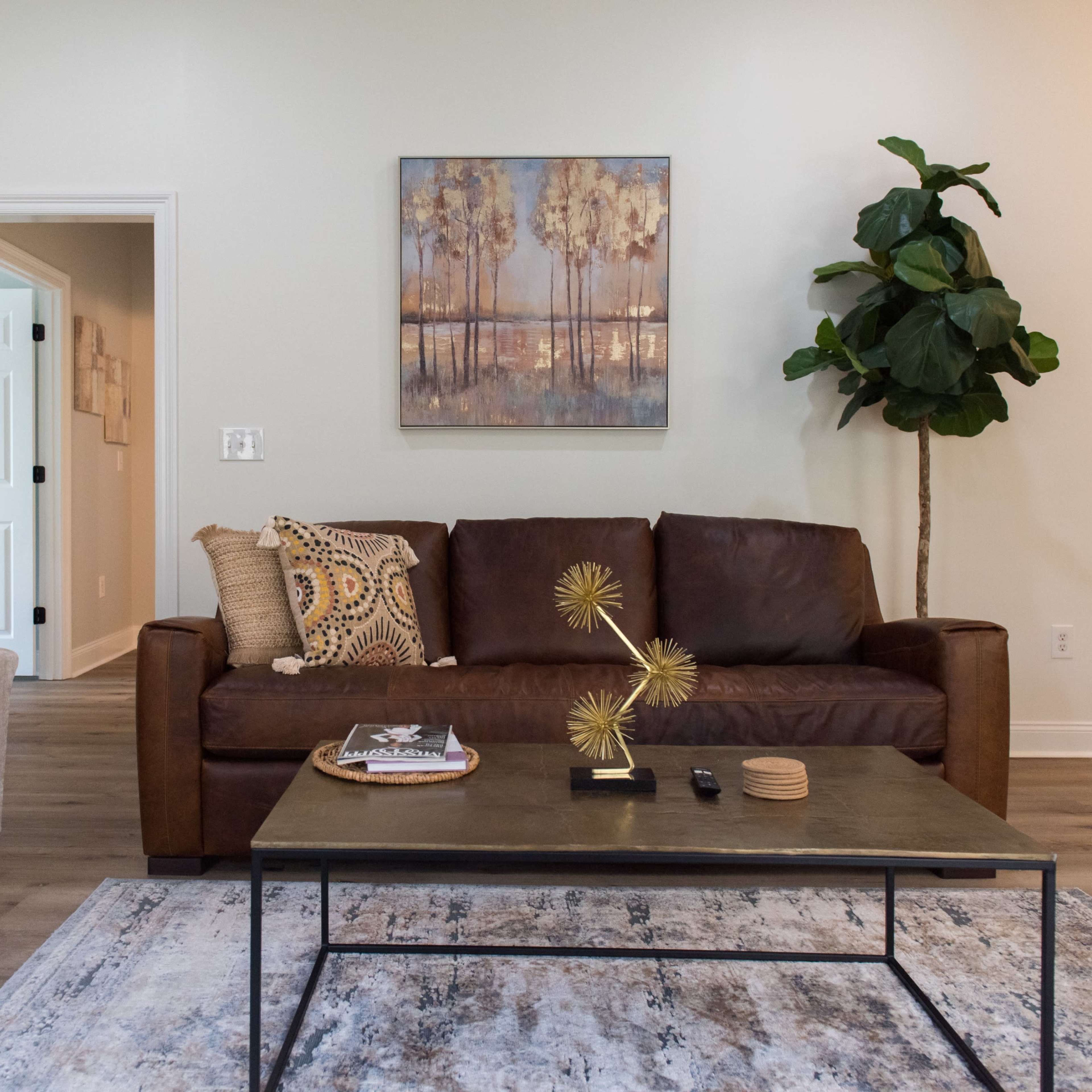 A brown leather sofa and a light gray armchair are arranged around a black coffee table on a patterned area rug in a well-lit living room featuring a wall painting and a potted plant.