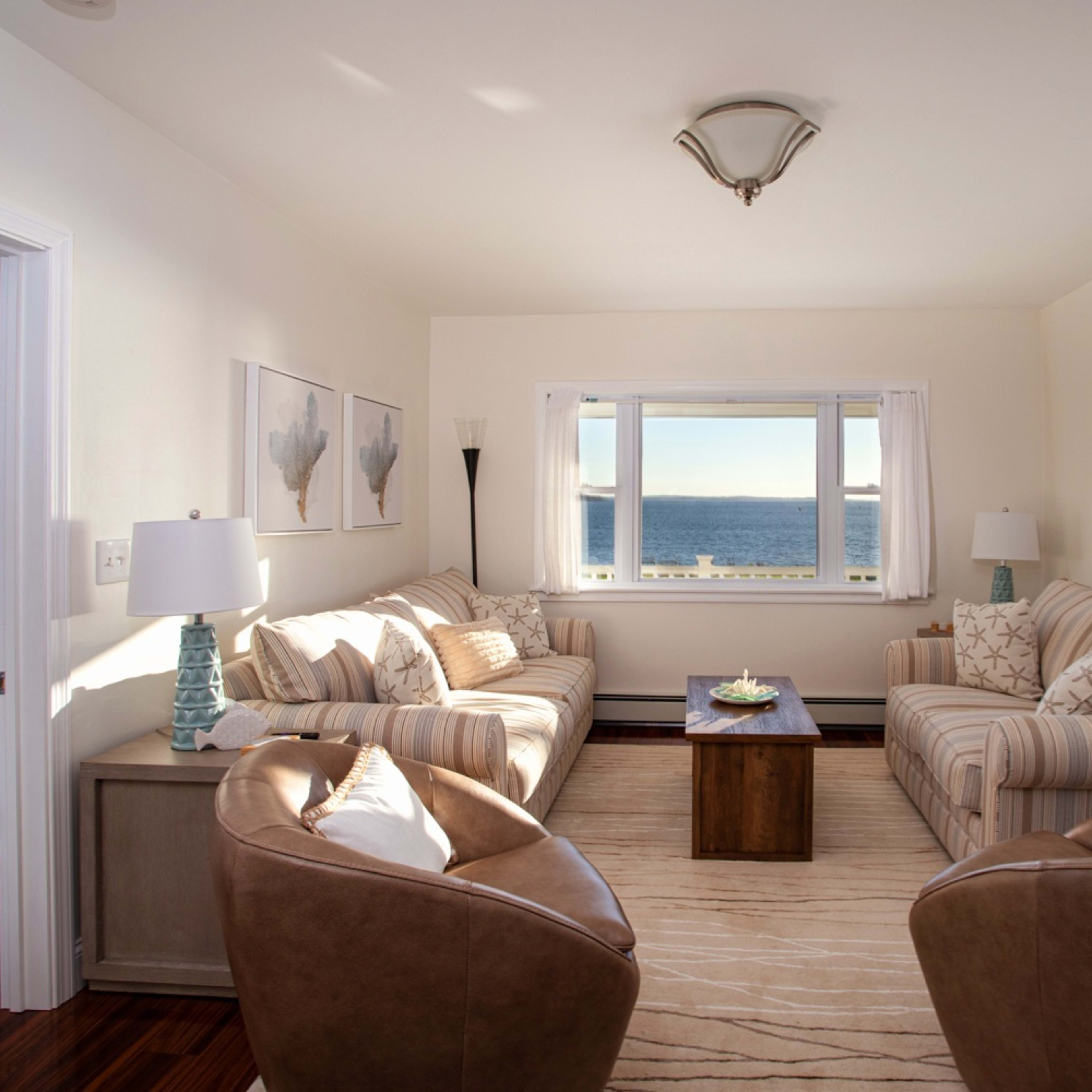 The room features a seating area with two couches and two chairs facing a coffee table, all set against a backdrop of large windows overlooking the ocean.