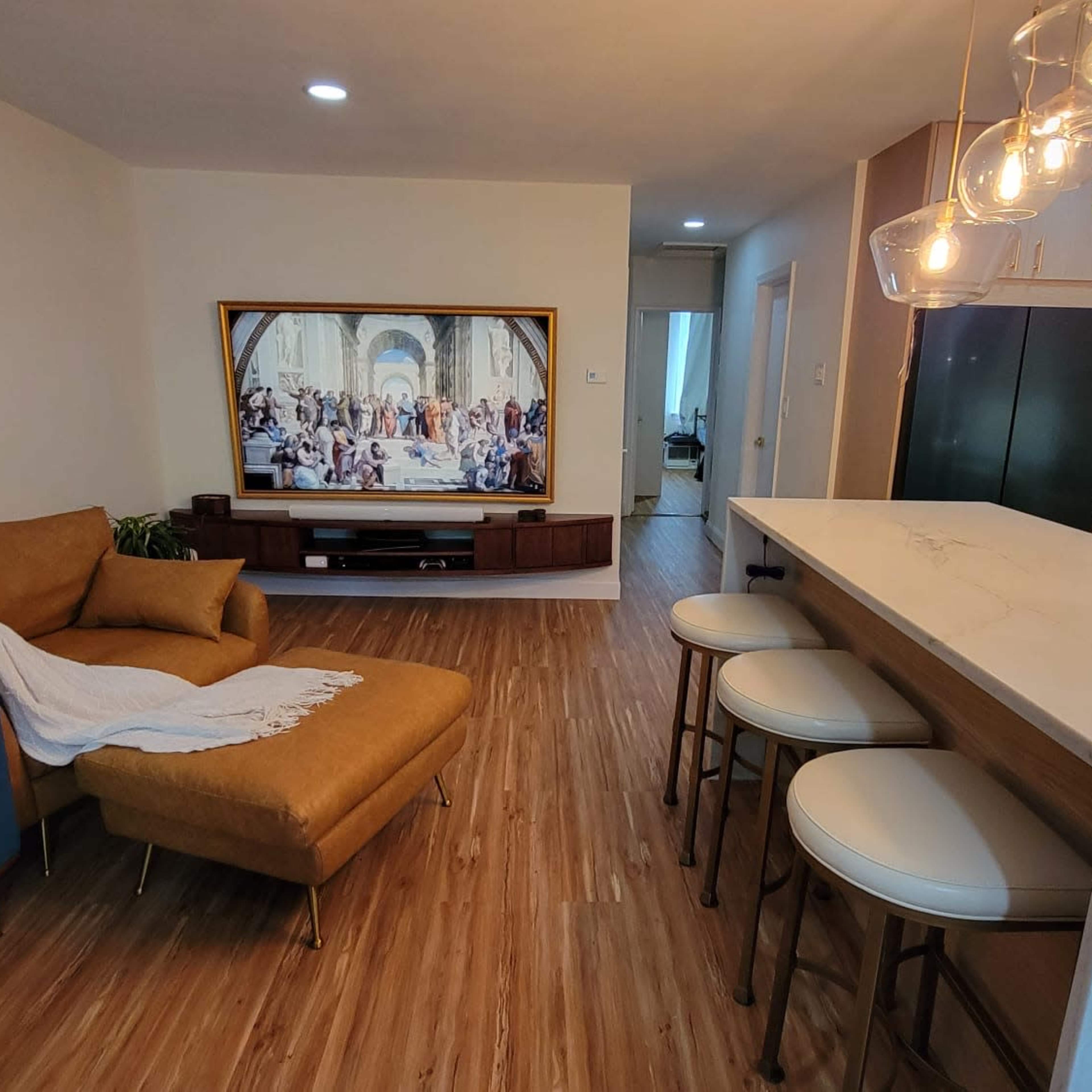 A modern living area featuring a blue sofa, a brown lounge chair, a marble-topped bar with white stools, and a large television displaying a classical painting.