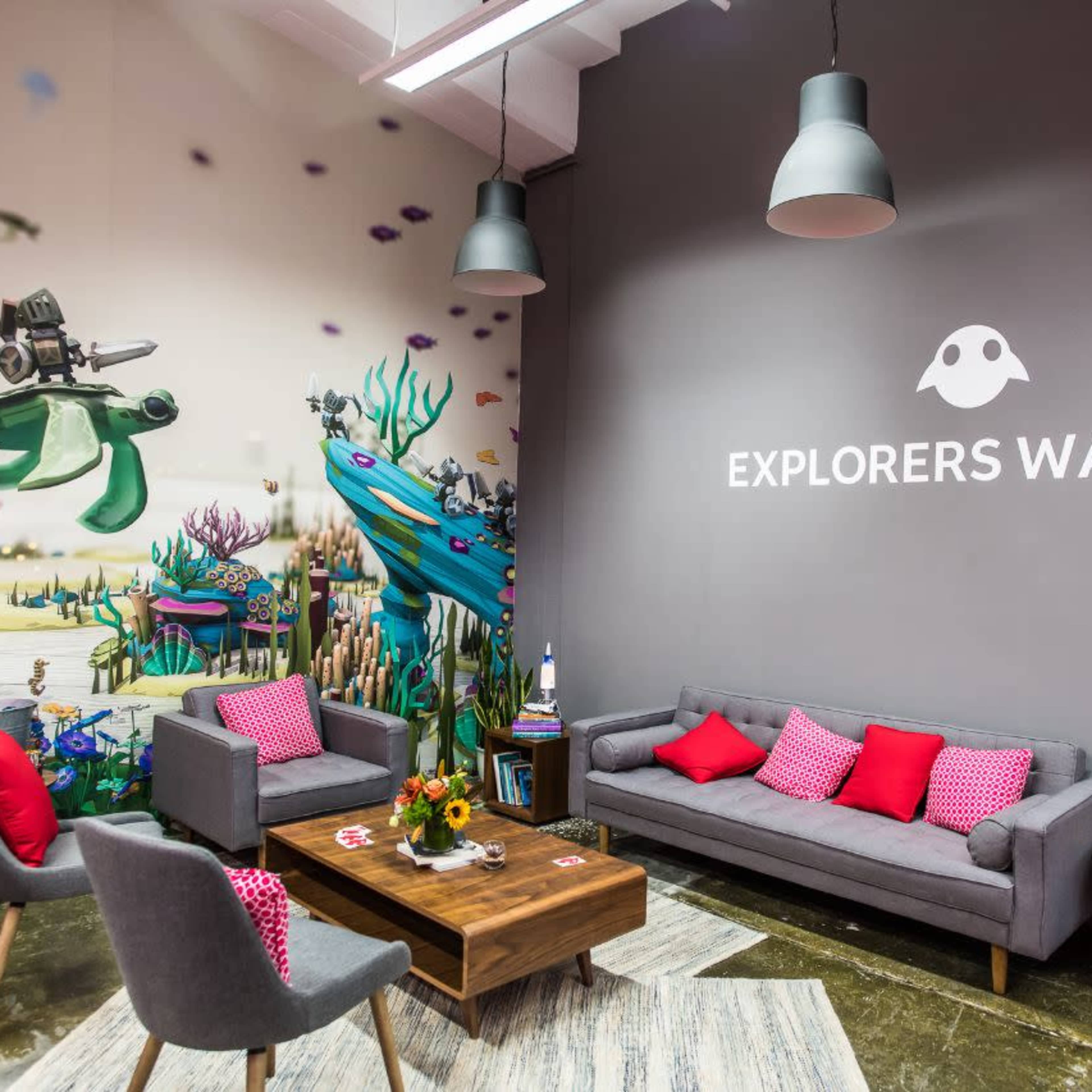 A modern lounge area with grey sofas, pink cushions, a wooden coffee table, and a colorful mural featuring underwater scenes on the wall.