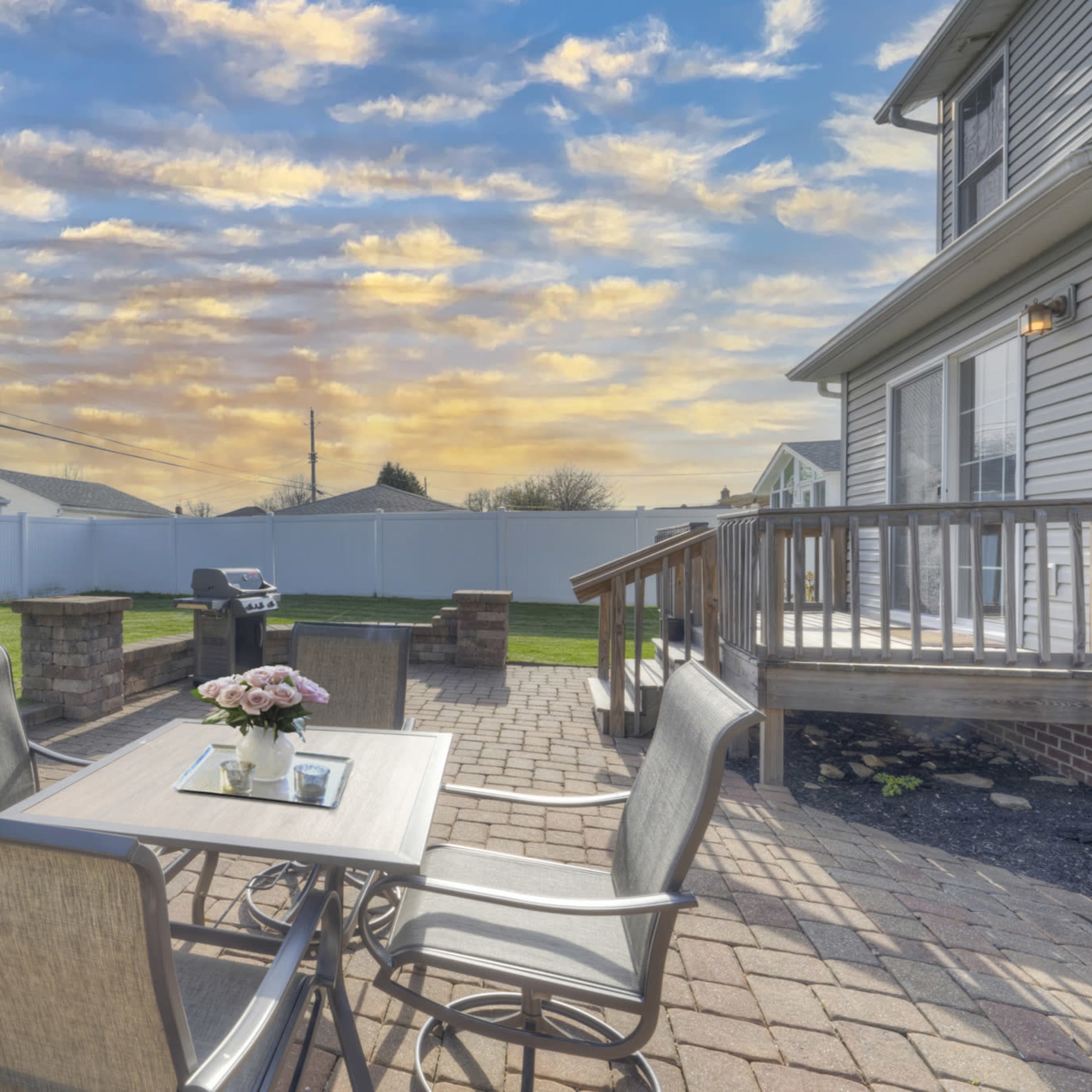 A patio area features a table with chairs, a grill, and a fence surrounding a grassy yard under a colorful sky.