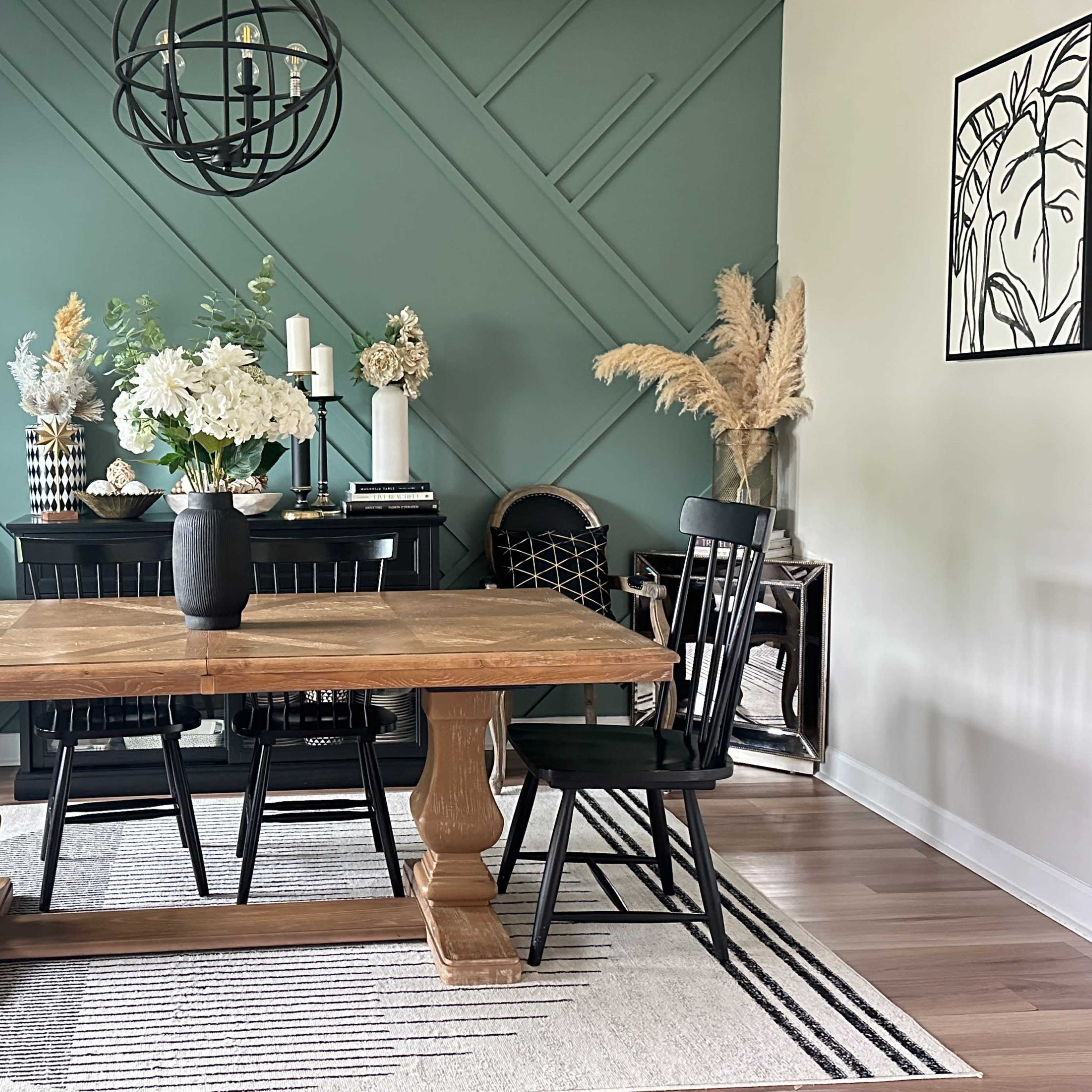 A dining area features a wooden table with black chairs, surrounded by decorative plants and framed artwork against a green wall.