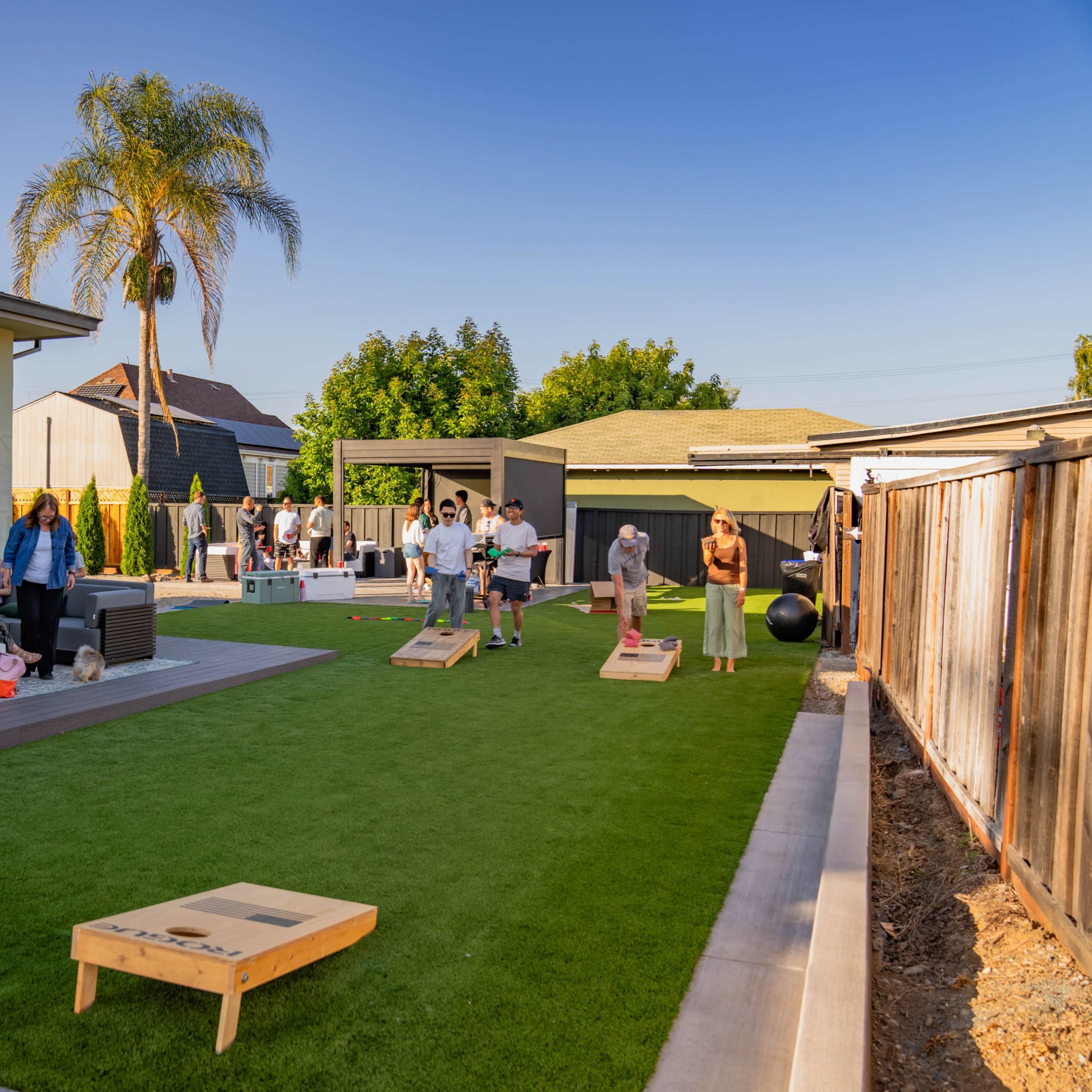 A group of people participates in lawn games in a spacious backyard surrounded by houses.