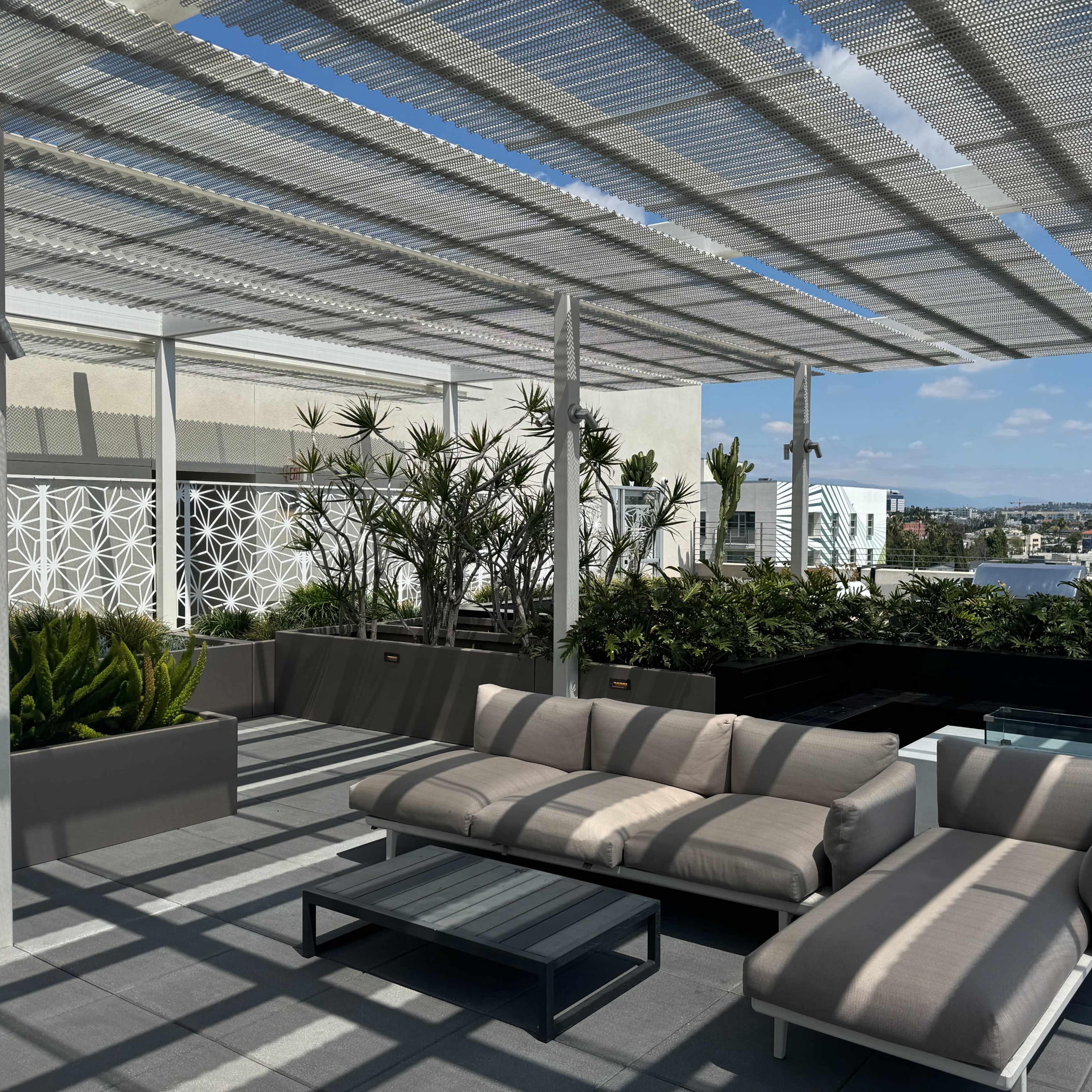 A modern rooftop lounge area features a couch and coffee table under a patterned pergola, surrounded by potted plants and a cityscape view.