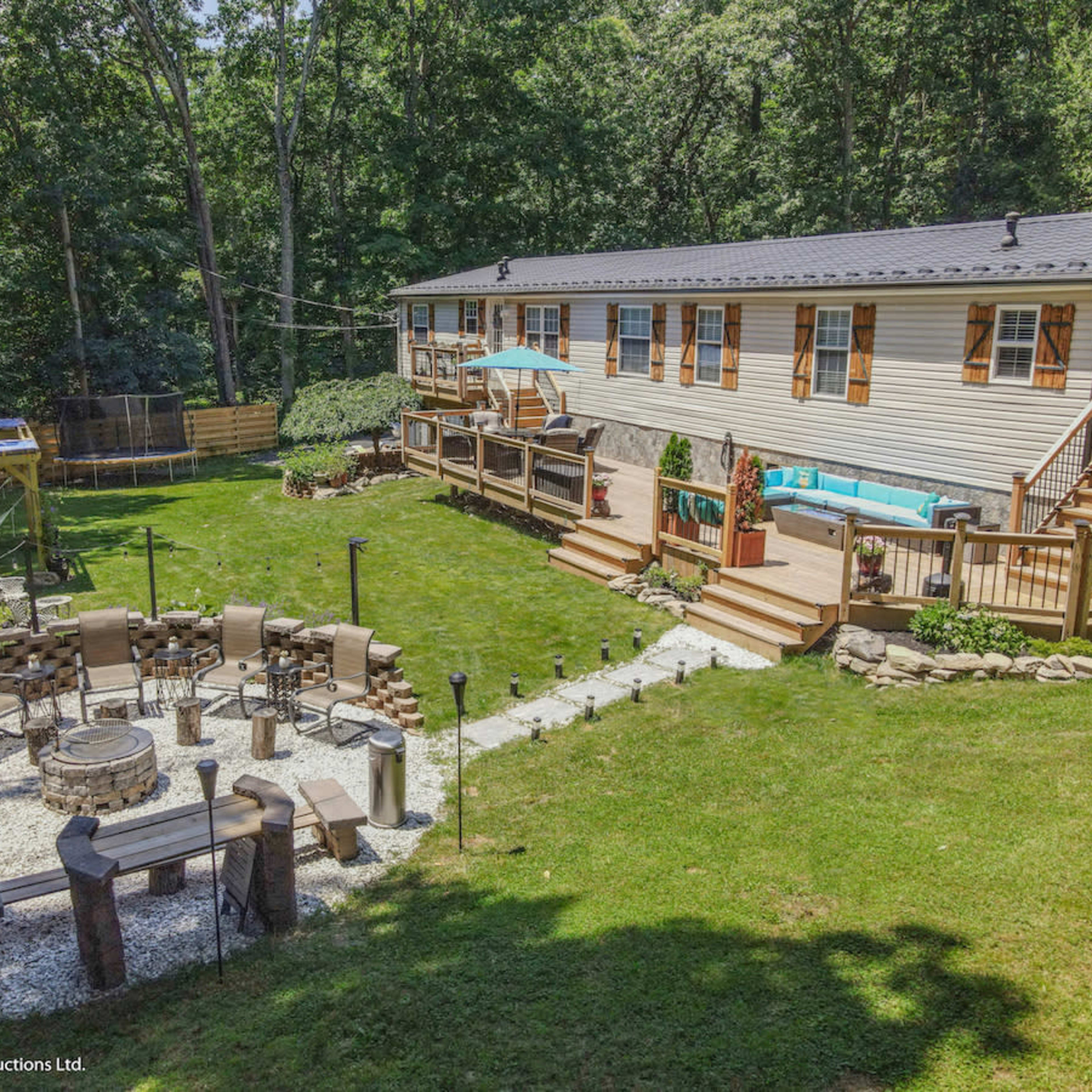 A landscaped backyard featuring a large wooden deck, a stone fire pit area surrounded by seating, and a well-maintained lawn, along with a house set against a wooded backdrop.
