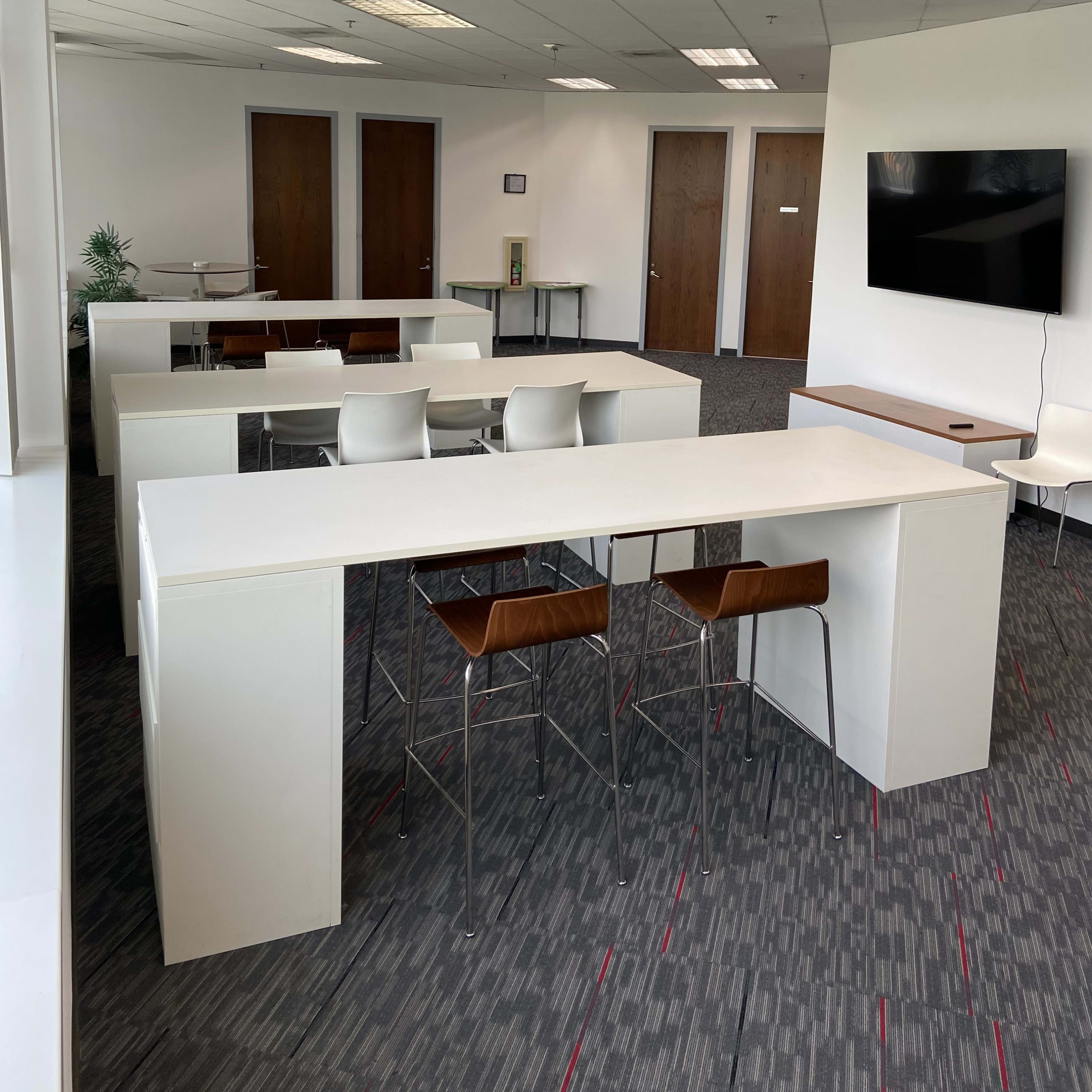 A modern office space with white tables and chairs arranged neatly, a large TV on the wall, and wooden doors in the background.