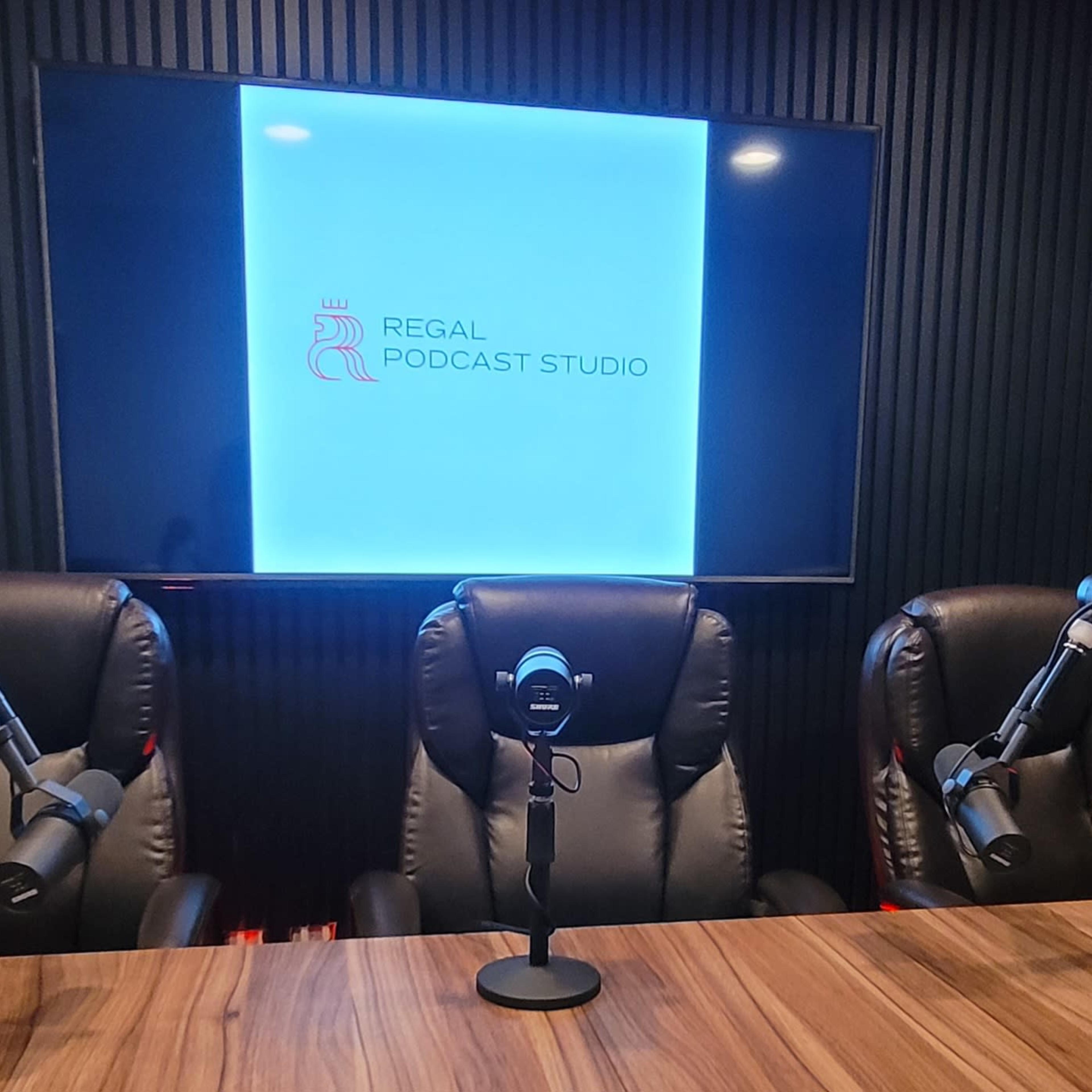 A podcast studio setup featuring three black chairs with microphone stands, a wooden table, and a large screen displaying the text "Regal Podcast Studio."
