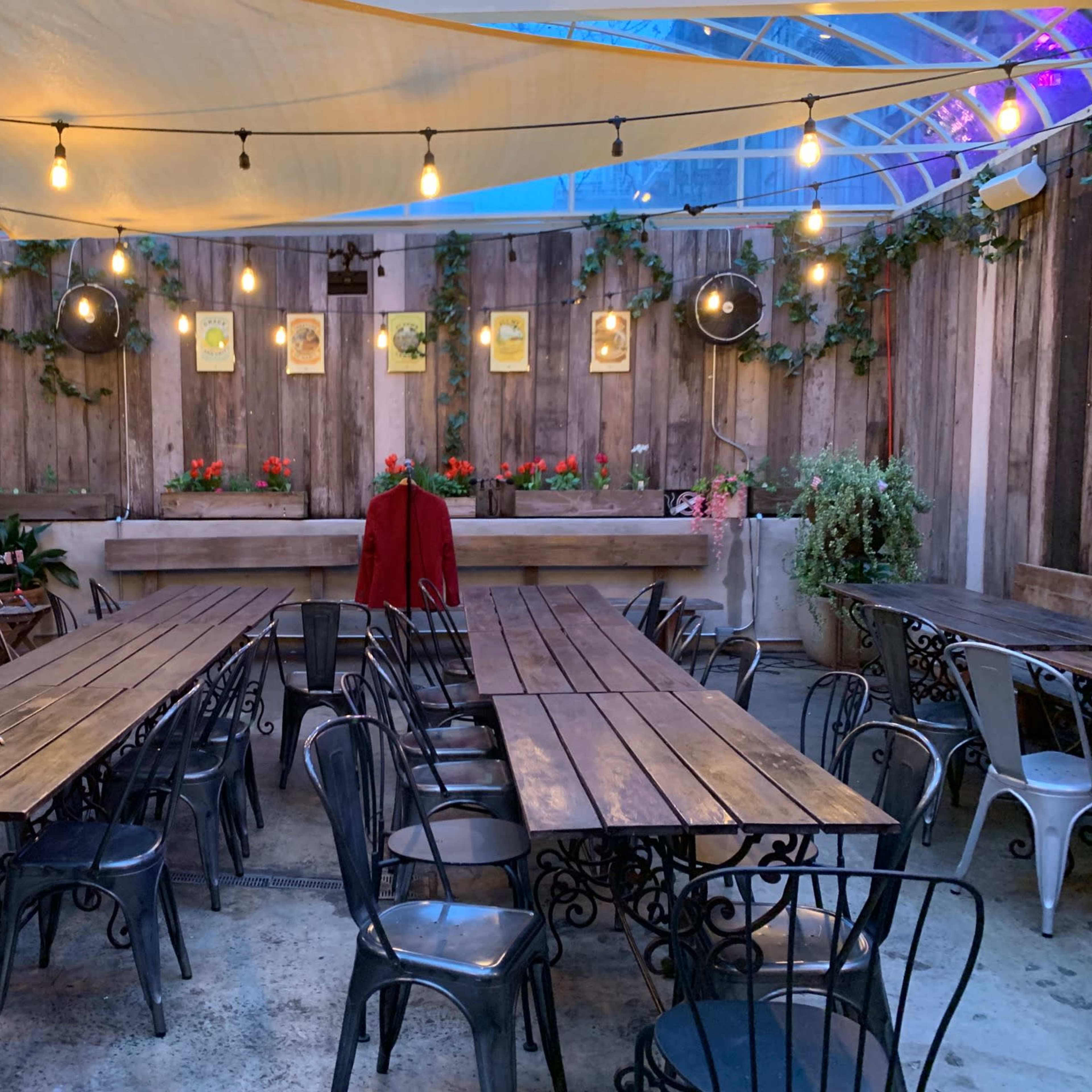 A spacious indoor dining area with wood tables, black metal chairs, string lights, and potted plants against a wooden wall.