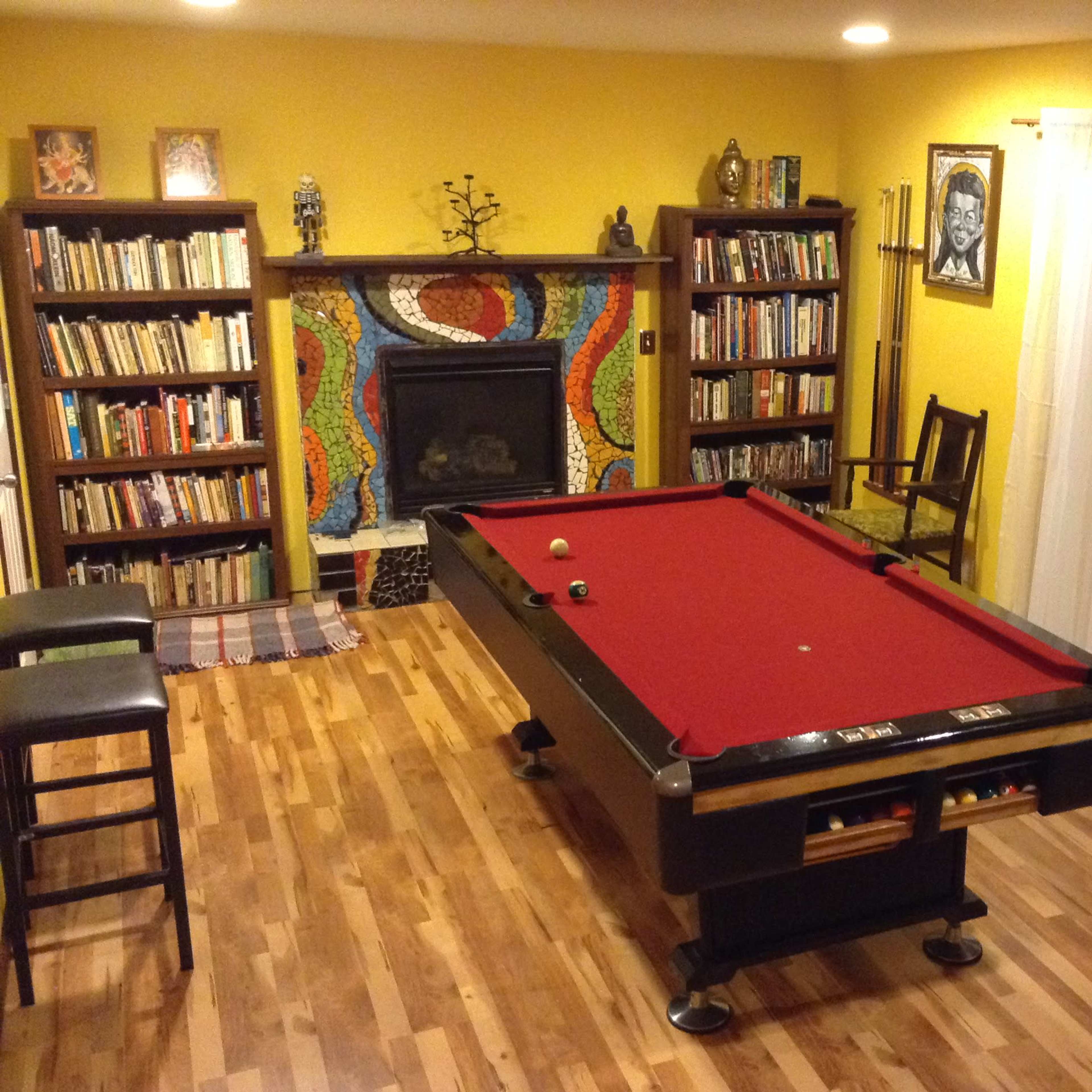 A game room with a pool table in the center, surrounded by bookshelves, a patterned fireplace, and a sliding glass door leading to an outside area.