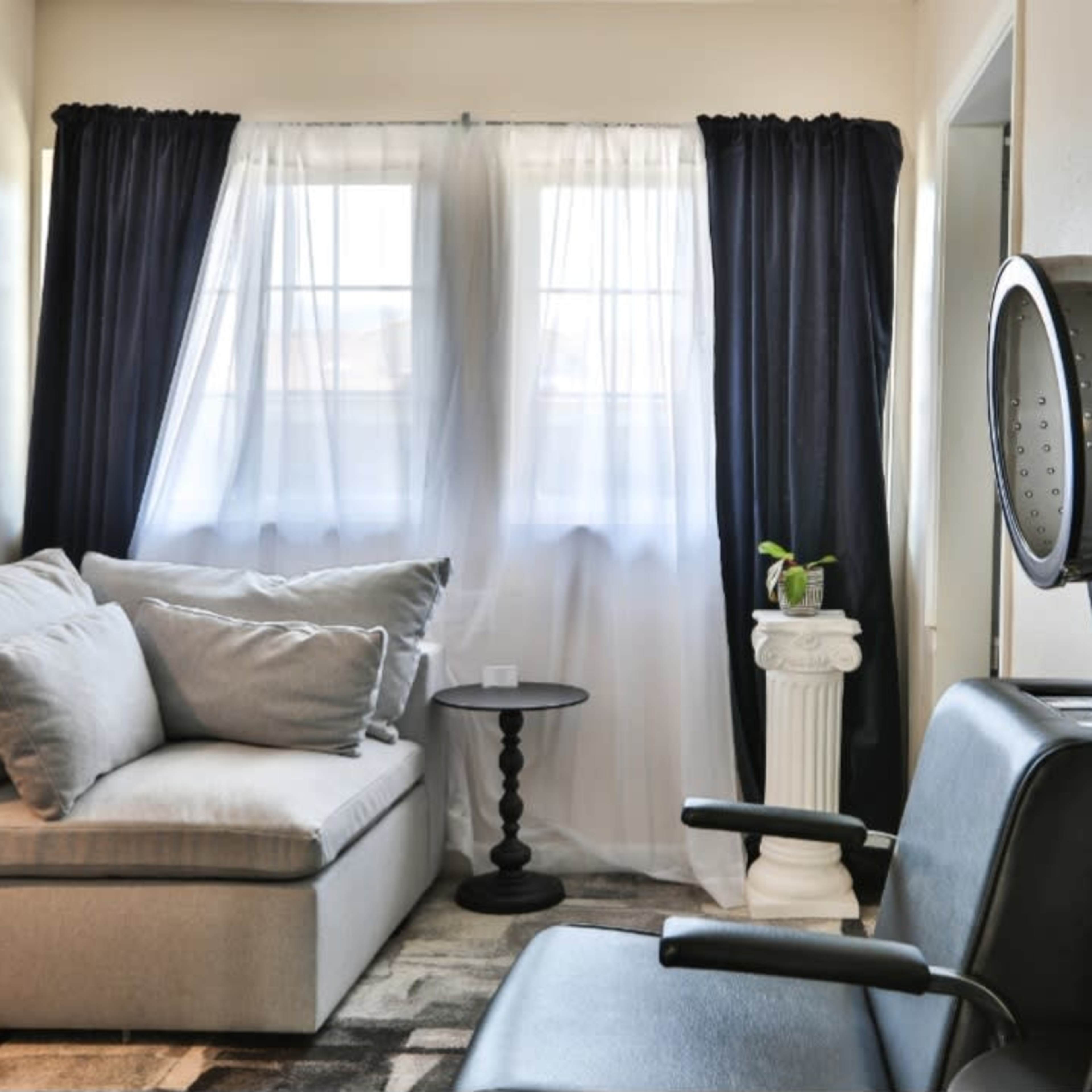 A cozy salon space with a black chair, a styling hood, a light-colored sofa, and large windows partially covered by sheer and dark curtains.