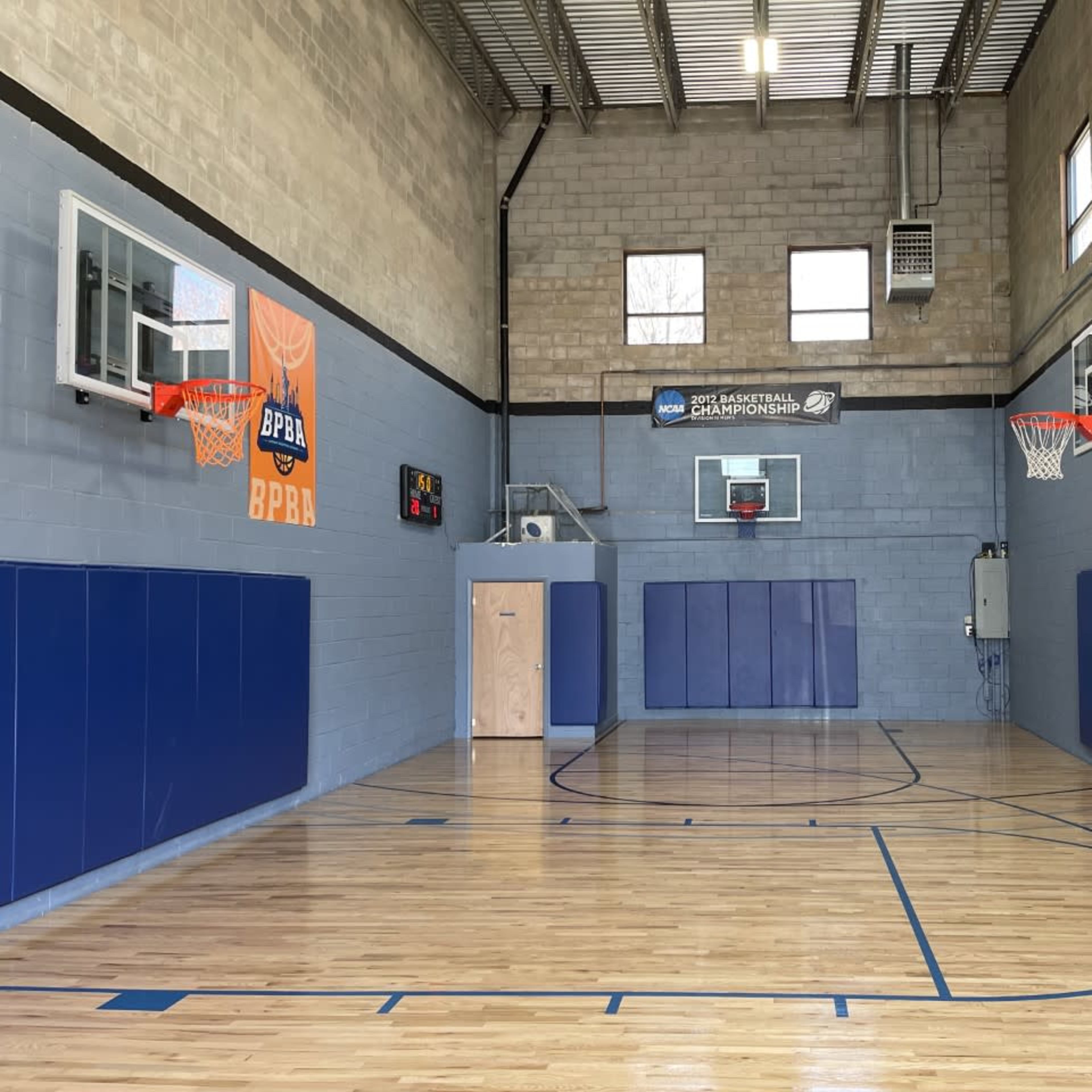 An indoor basketball court featuring two hoops, blue wall padding, and an American flag on the wall.