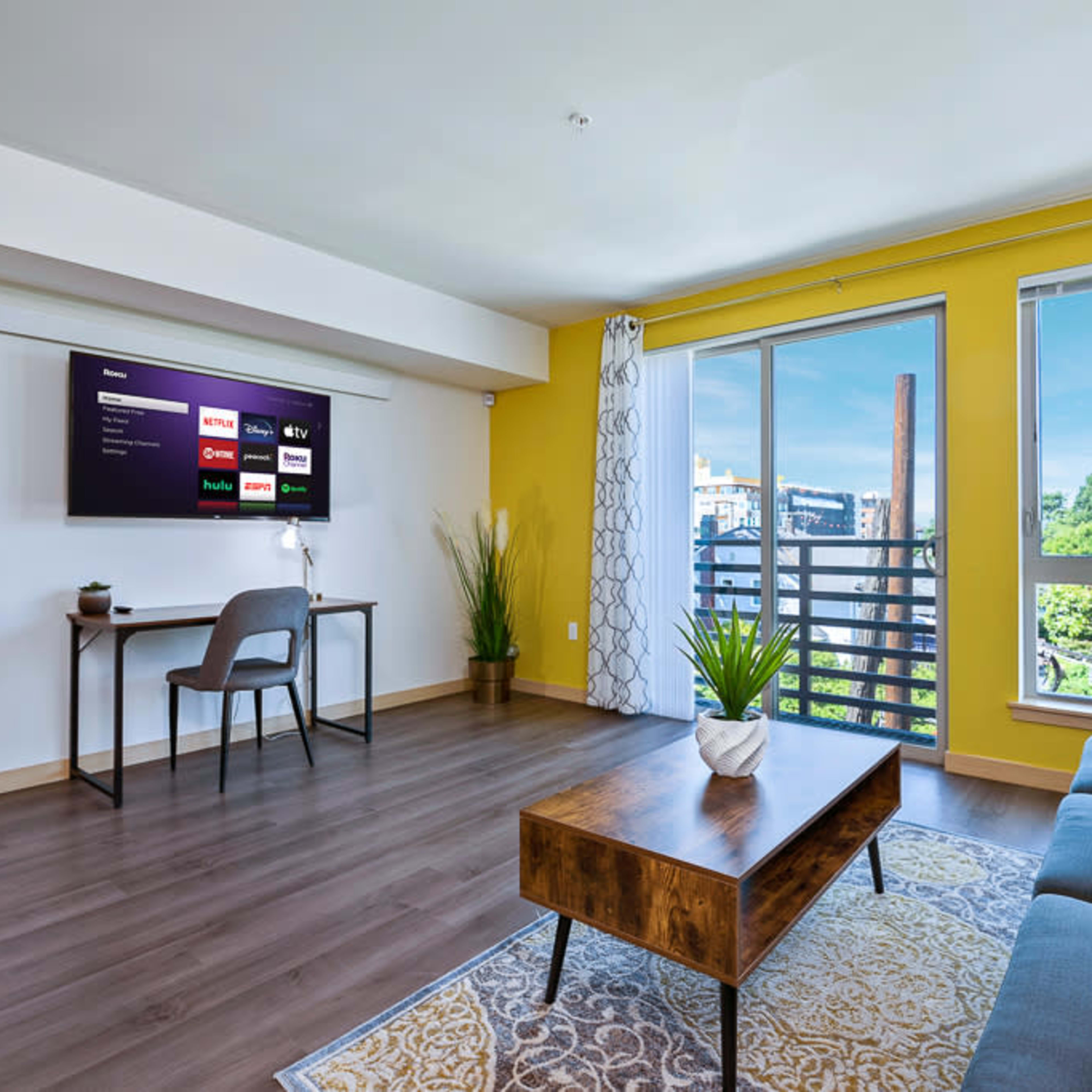 A modern living room features a yellow accent wall, a comfortable gray sofa, a wooden coffee table, a small desk with a chair, and a large window with a balcony view.