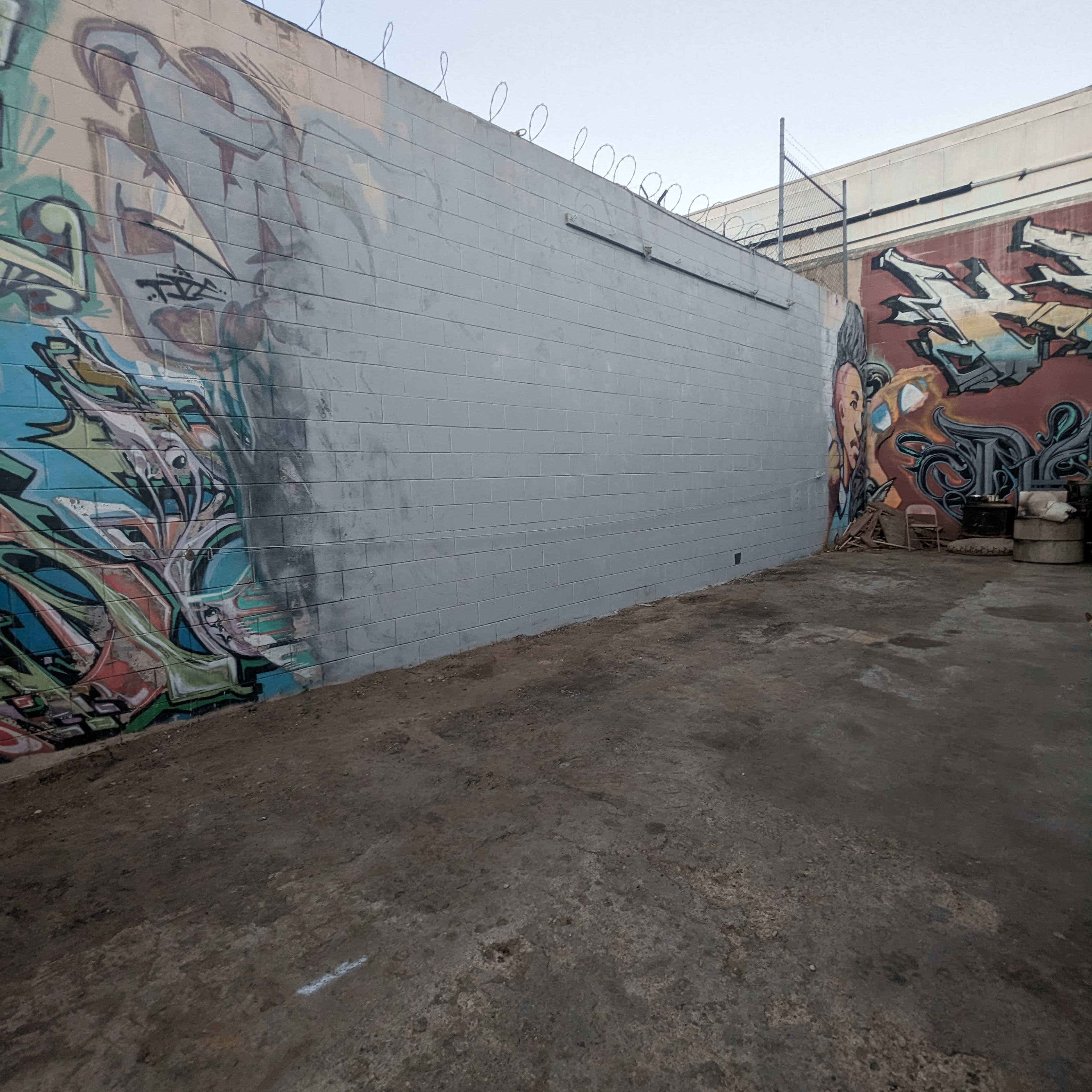 A concrete wall beside a colorful mural, with barbed wire on top, in a gravel area.