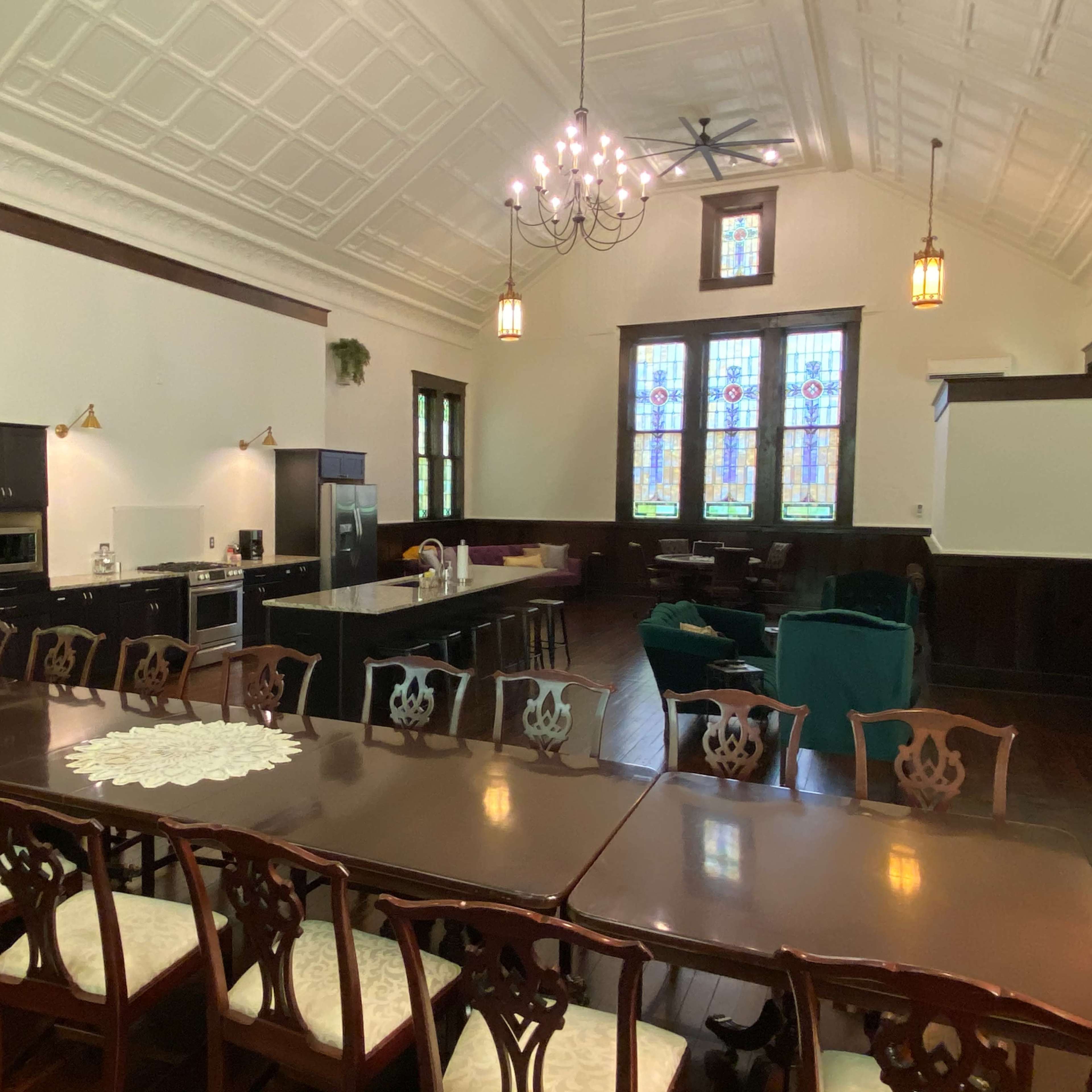 A spacious room with a long dining table, a modern kitchen area, and large stained glass windows.