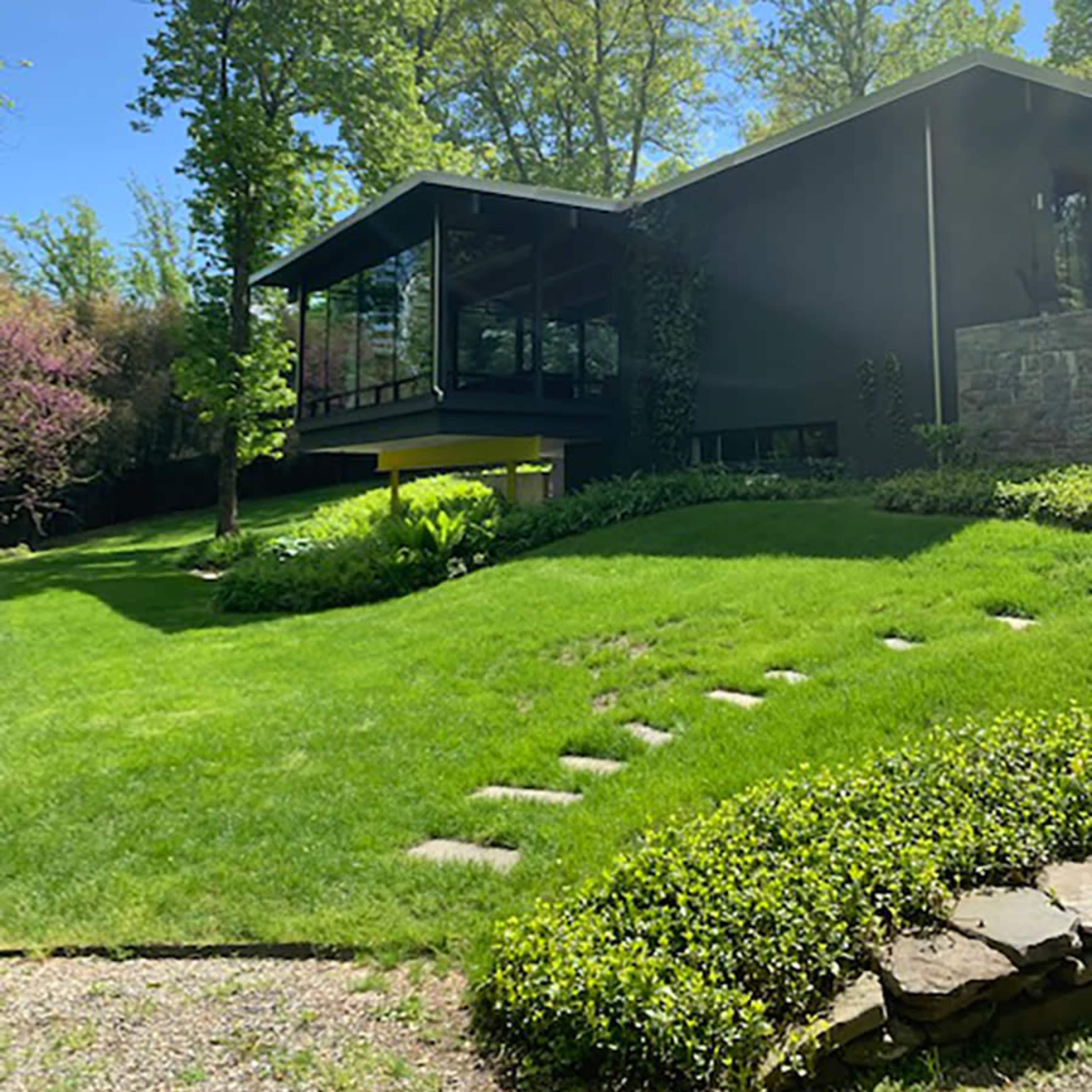 A modern house with large glass windows is set on a gently sloping green lawn surrounded by trees.