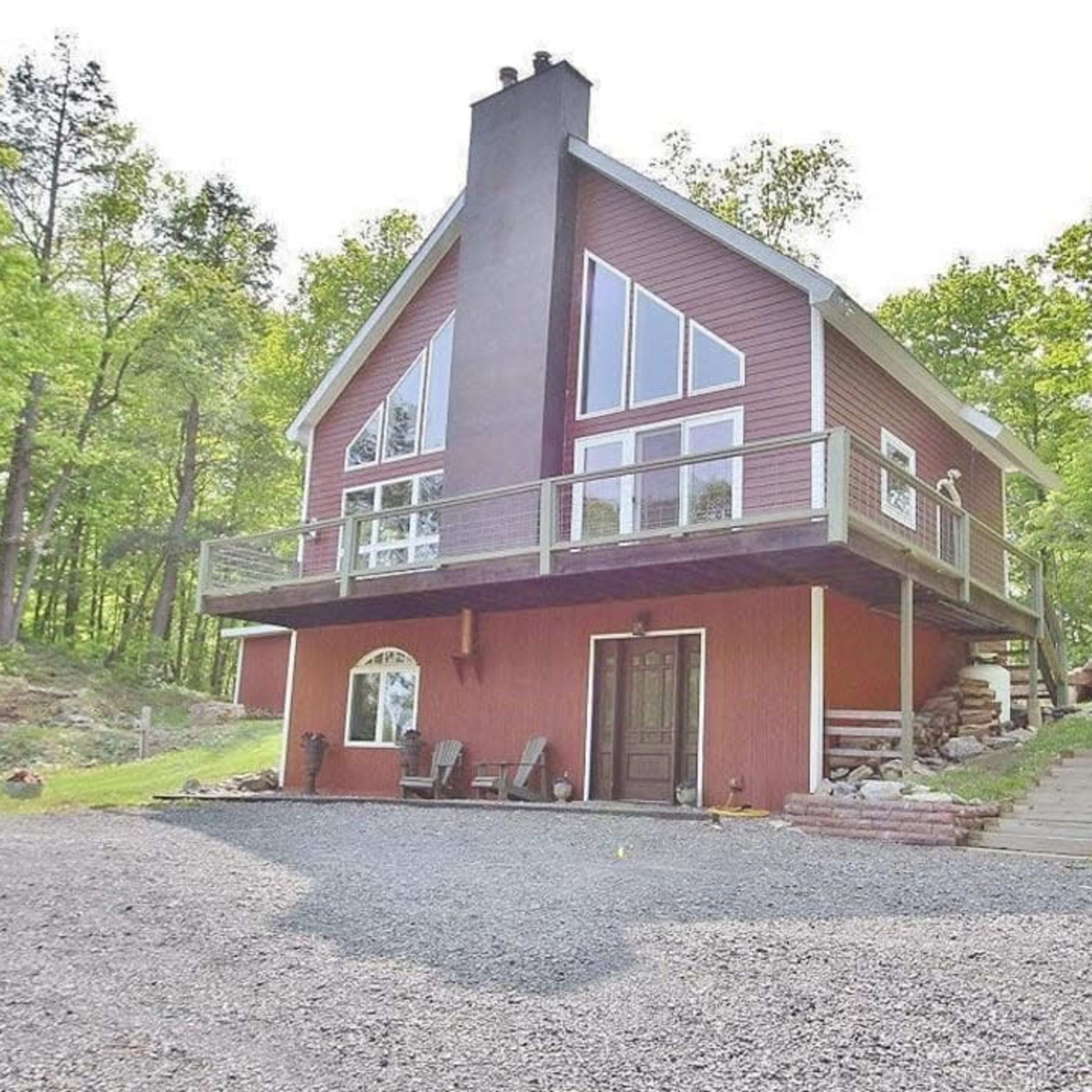 A two-story red house with large windows and a balcony is situated on a gravel driveway surrounded by trees.